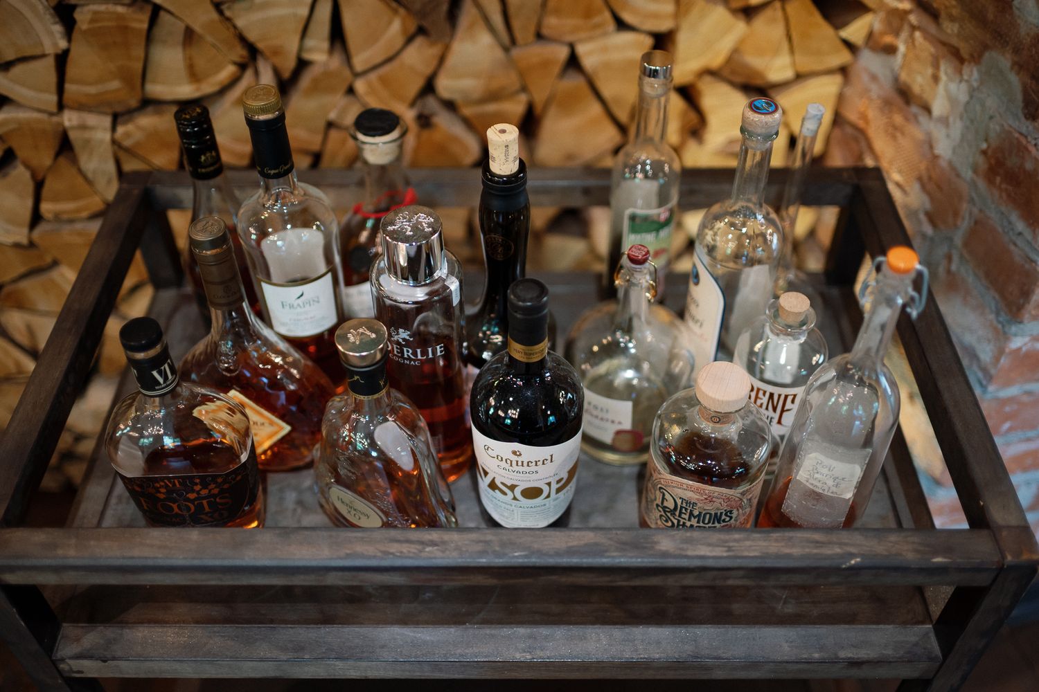 A wooden tray filled with various liquor bottles displayed against a rustic firewood wall backdrop.