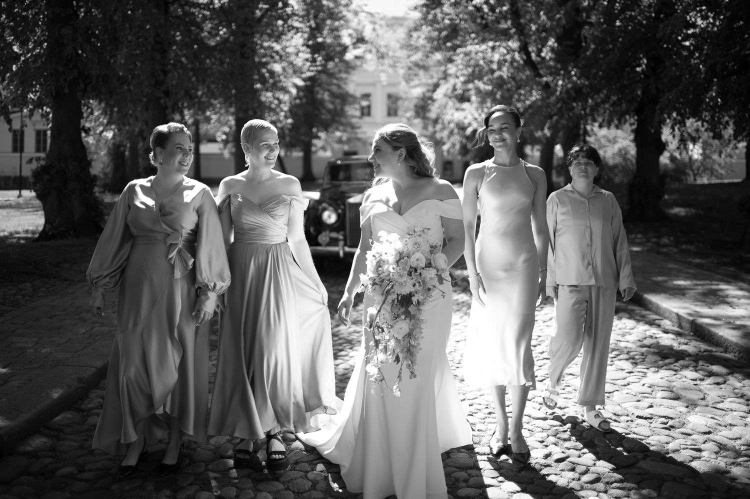 Black and white photo of a bride holding a bouquet with four bridesmaids in elegant dresses outdoors.