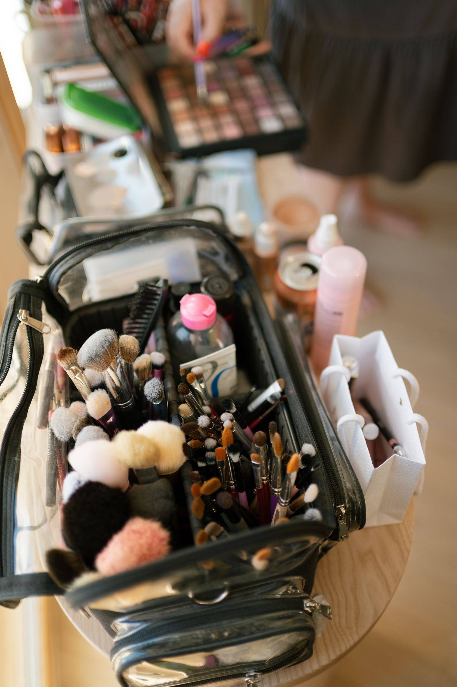 Open makeup artist case filled with brushes and cosmetics on a table, with beauty products and palette visible in background.
