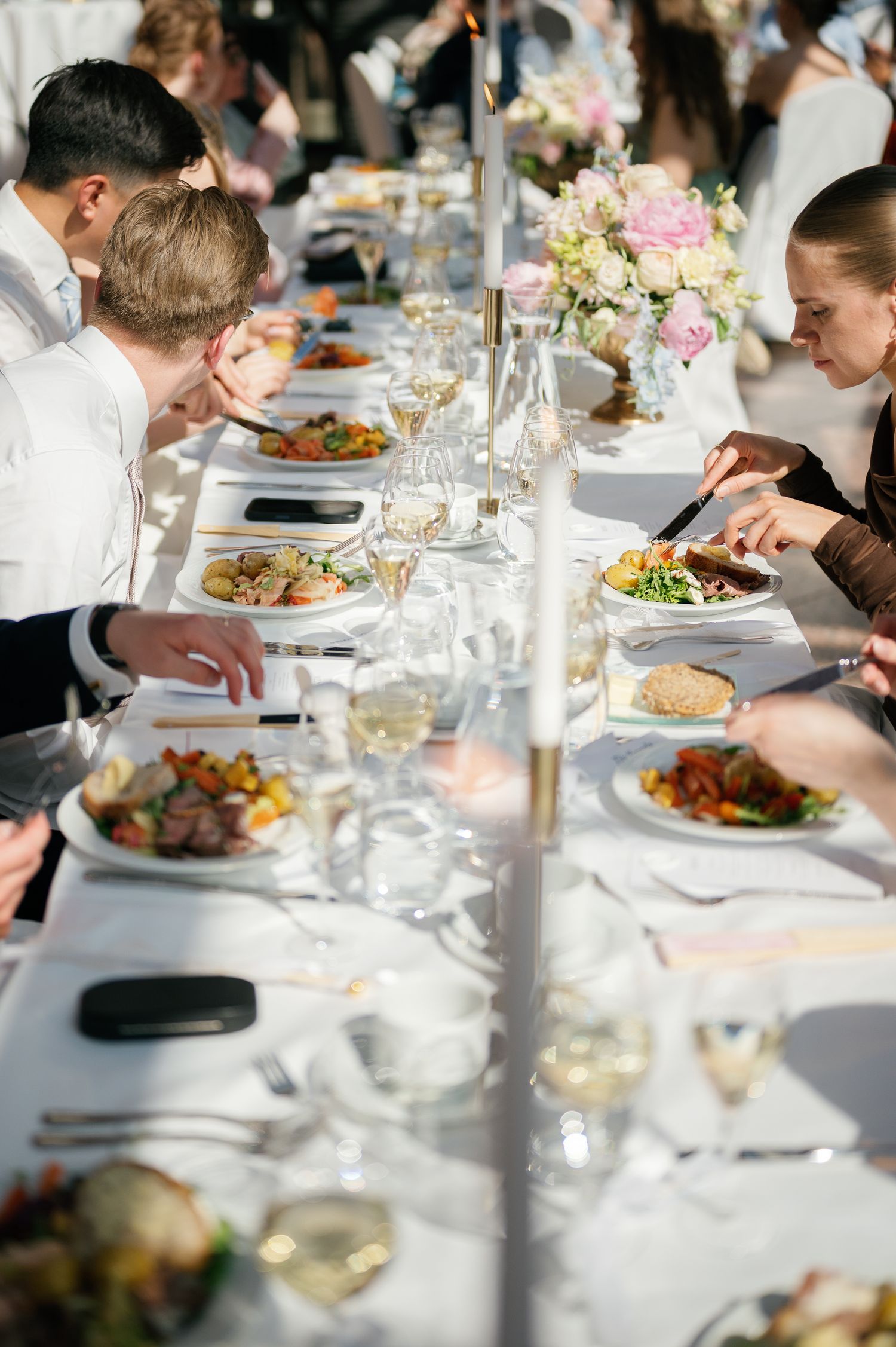 Guests dining at a long elegant banquet table with white linens, floral centerpieces, and plated meals during a formal event.