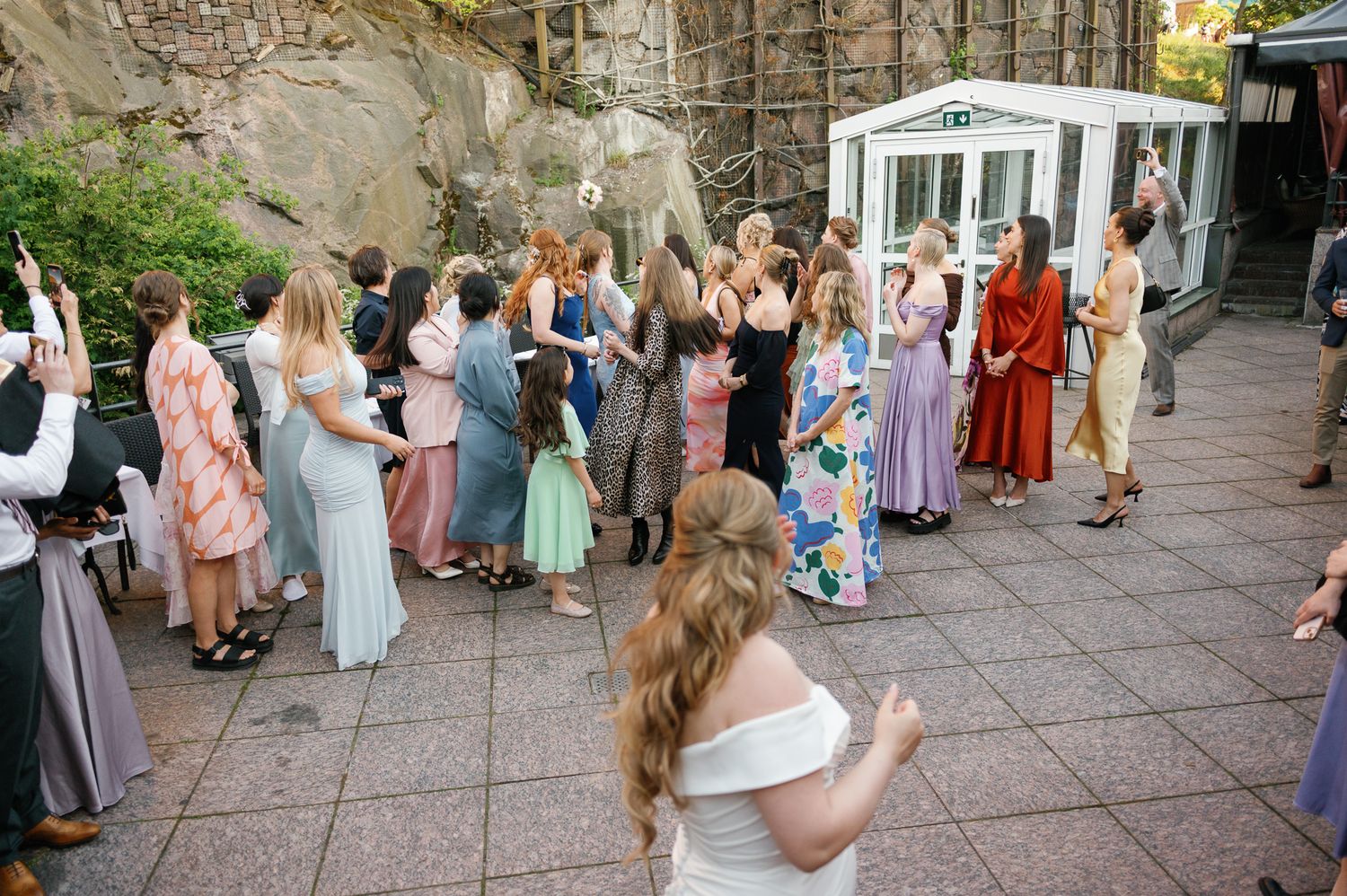 Wedding guests in colorful dresses gather outdoors on a patio near a stone wall during a bouquet toss.