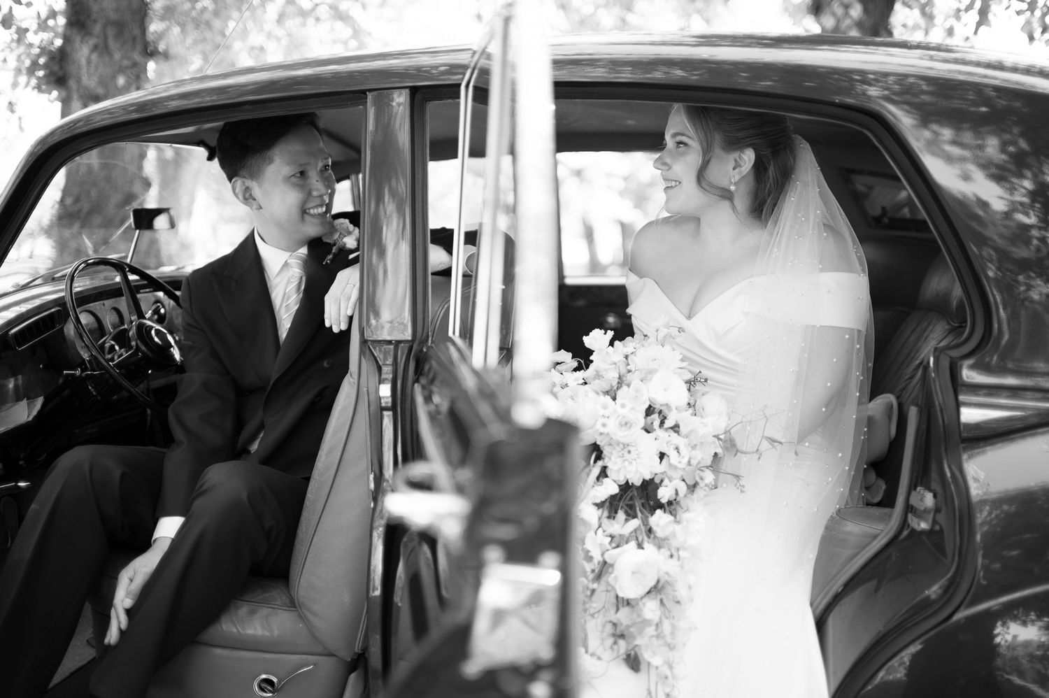 Black and white photo of a bride holding flowers and groom sitting in a vintage classic car on their wedding day.