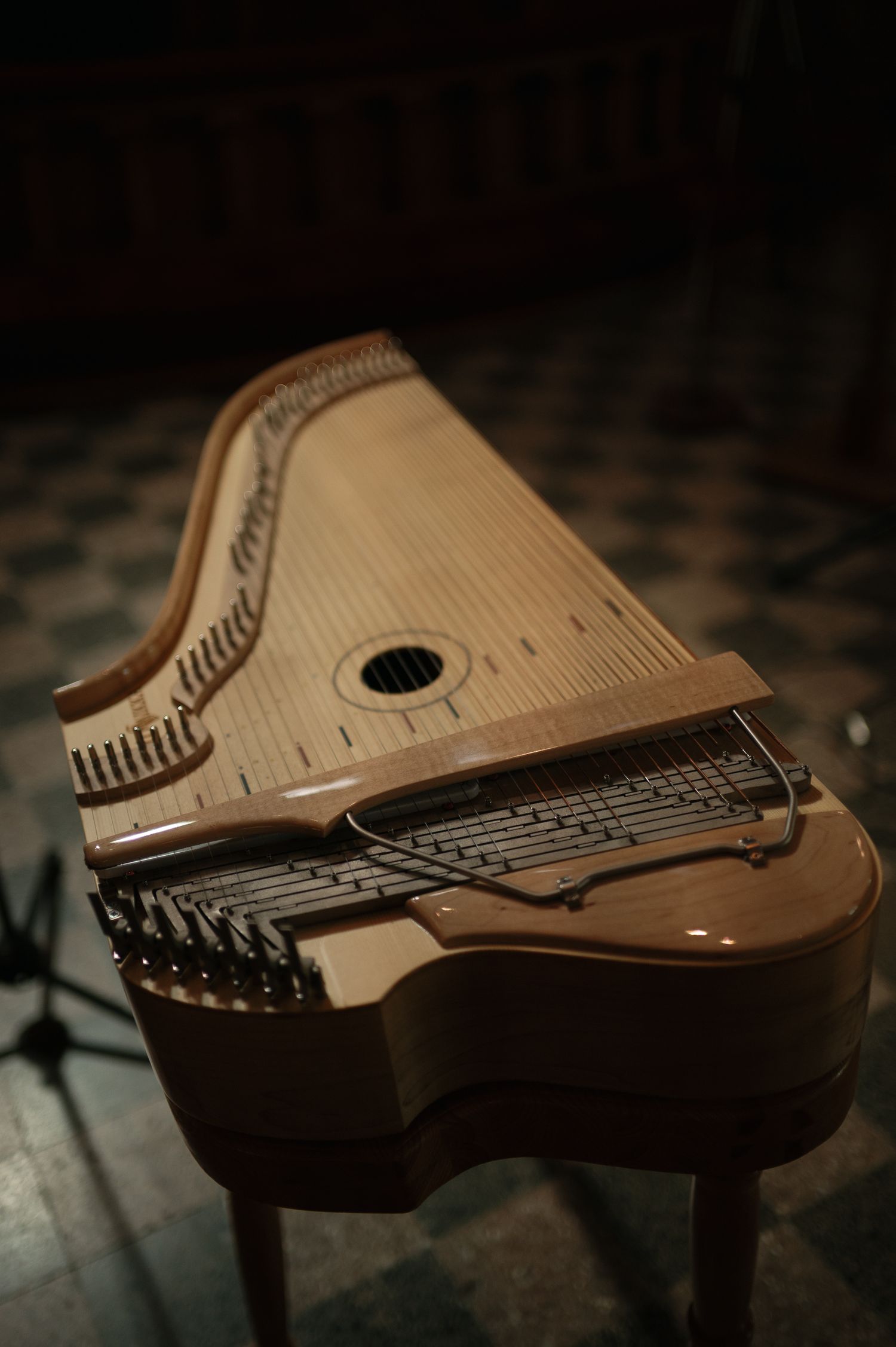 A wooden harpsichord with visible strings and keys displayed on a tiled floor in dim lighting.