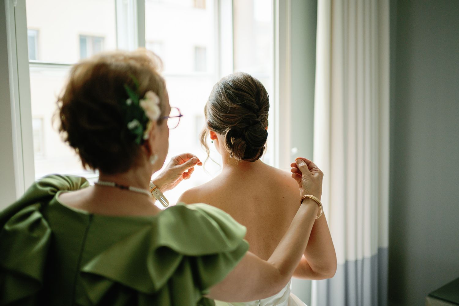 Mother helping bride with dress near window, both seen from behind, bride in white gown, mother in green ruffled top.