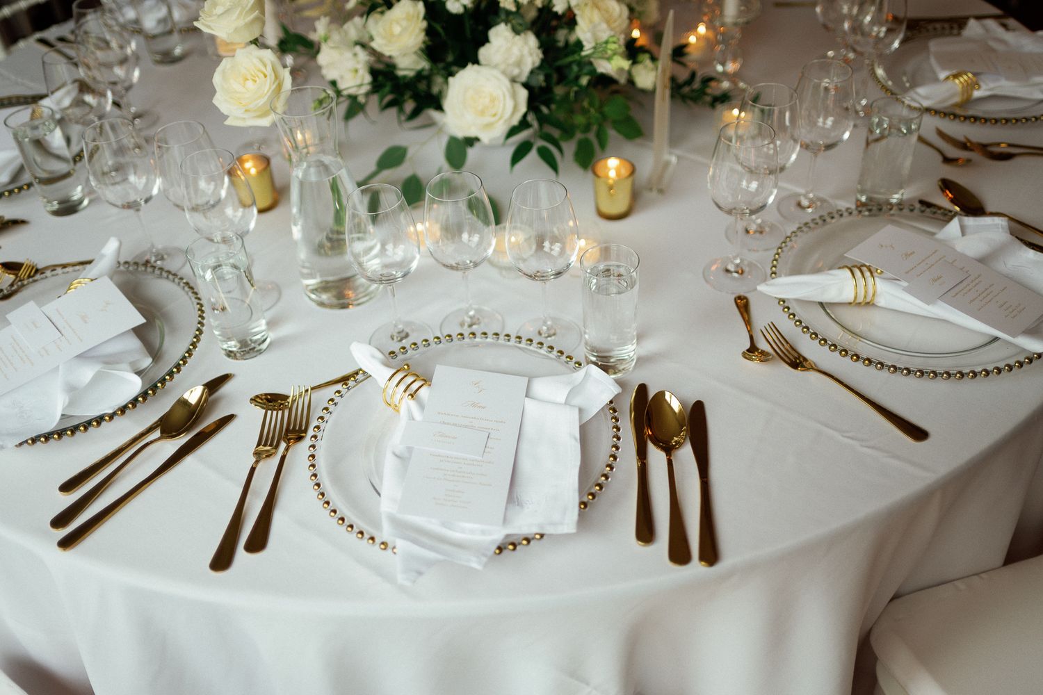 Elegant wedding reception table with gold cutlery, beaded charger plates, white floral centerpiece, and crystal glassware on white linen.