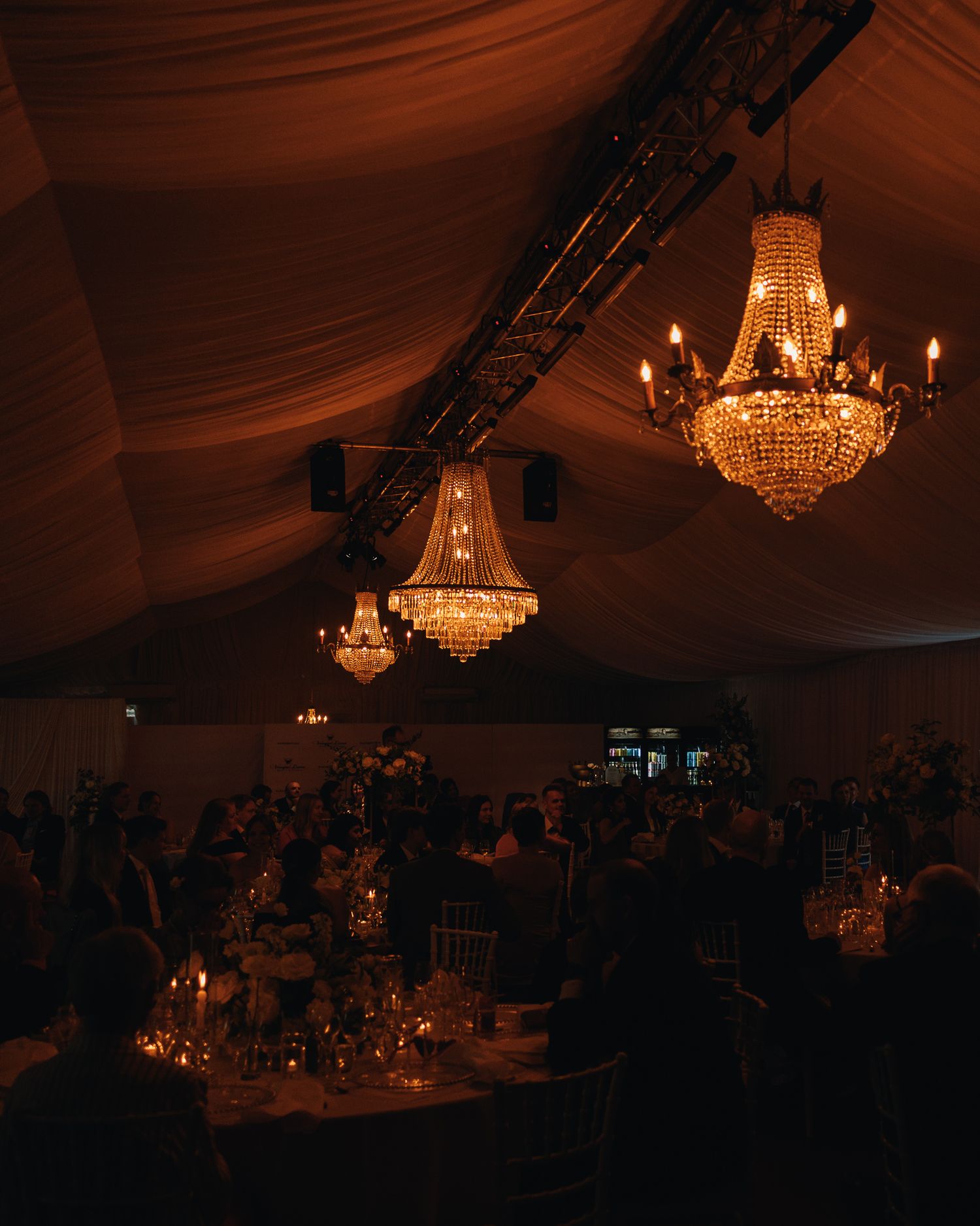 Elegant wedding reception tent with crystal chandeliers casting warm golden light over candlelit tables and guests.