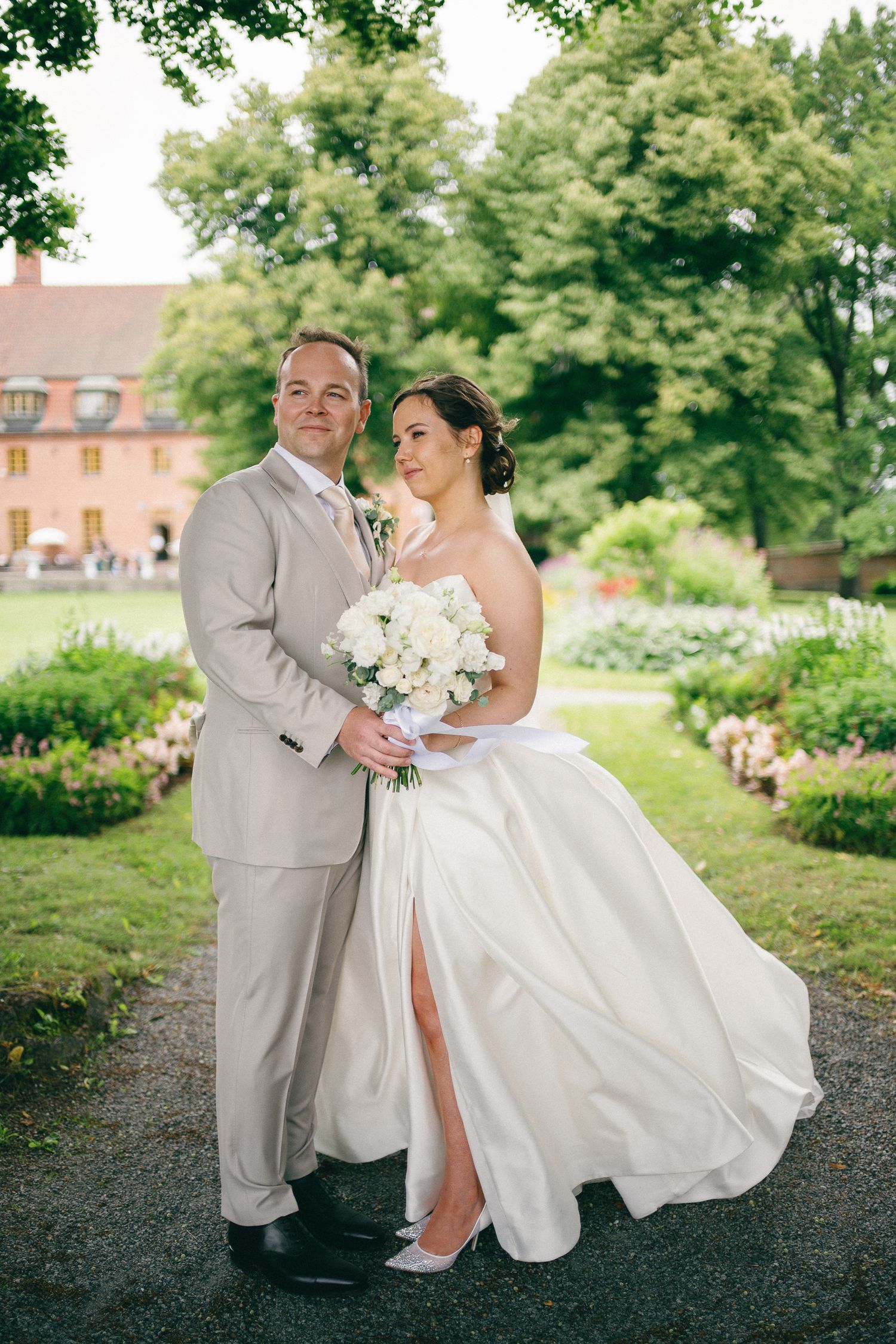 Bride and groom pose in garden, she holds white bouquet in wedding gown, he wears beige suit, lush greenery behind.