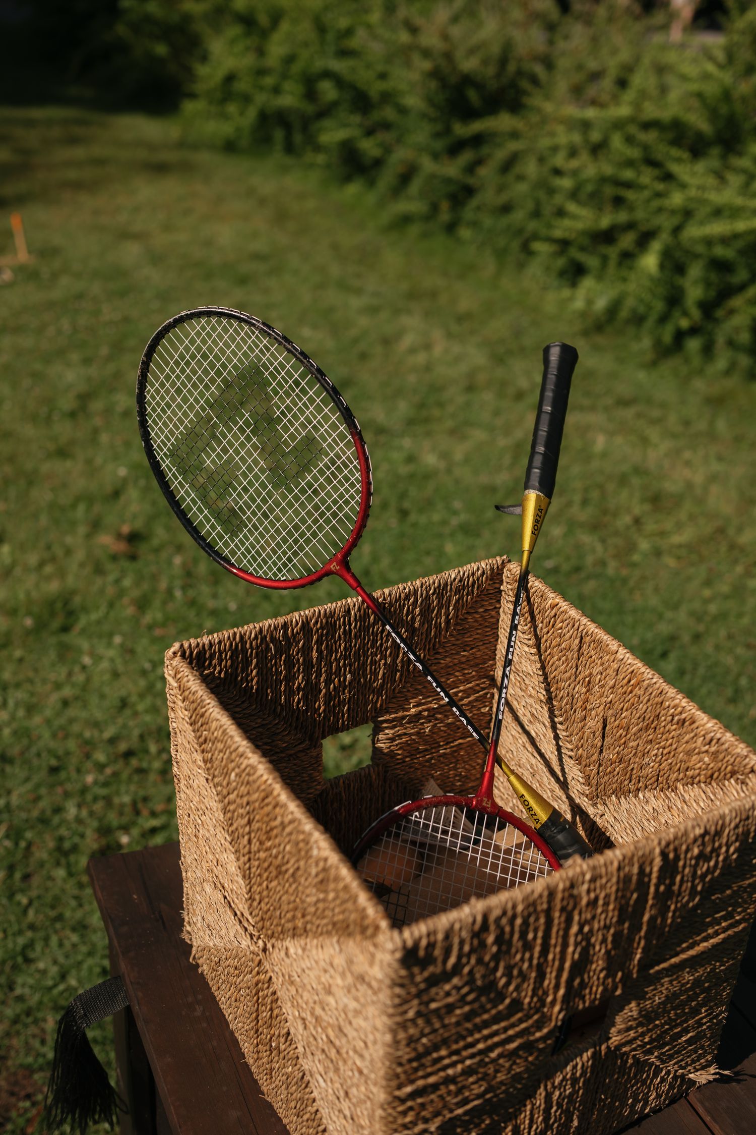 Two red and yellow badminton rackets stored in a woven wicker basket on green grass outdoors.