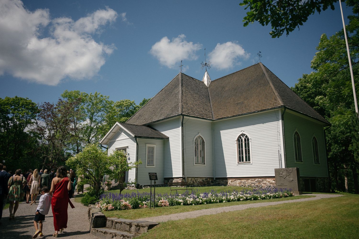 A charming white Nordic church with a gray roof surrounded by lush green trees and a flower garden on a sunny day.