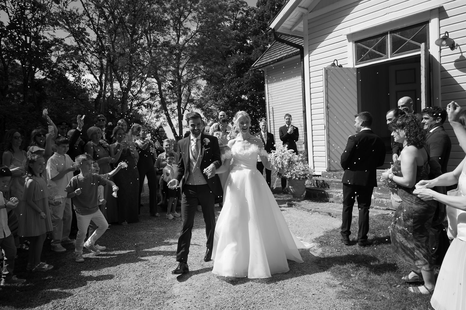Black and white photo of a bride and groom walking through a confetti celebration with cheering guests outside a white building.
