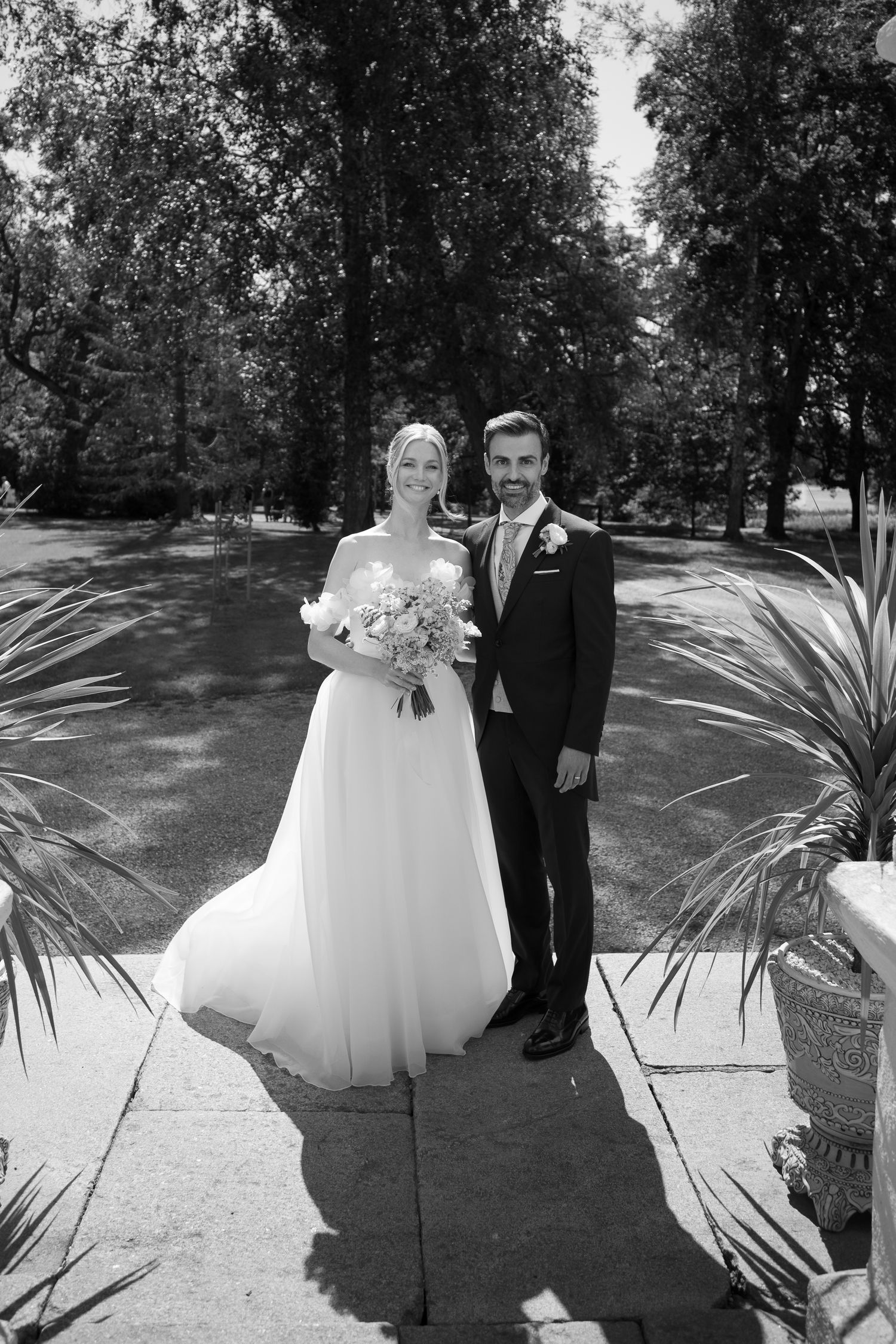 Black and white photo of a bride and groom posing outdoors on a sunny day, surrounded by lush trees and tropical plants.