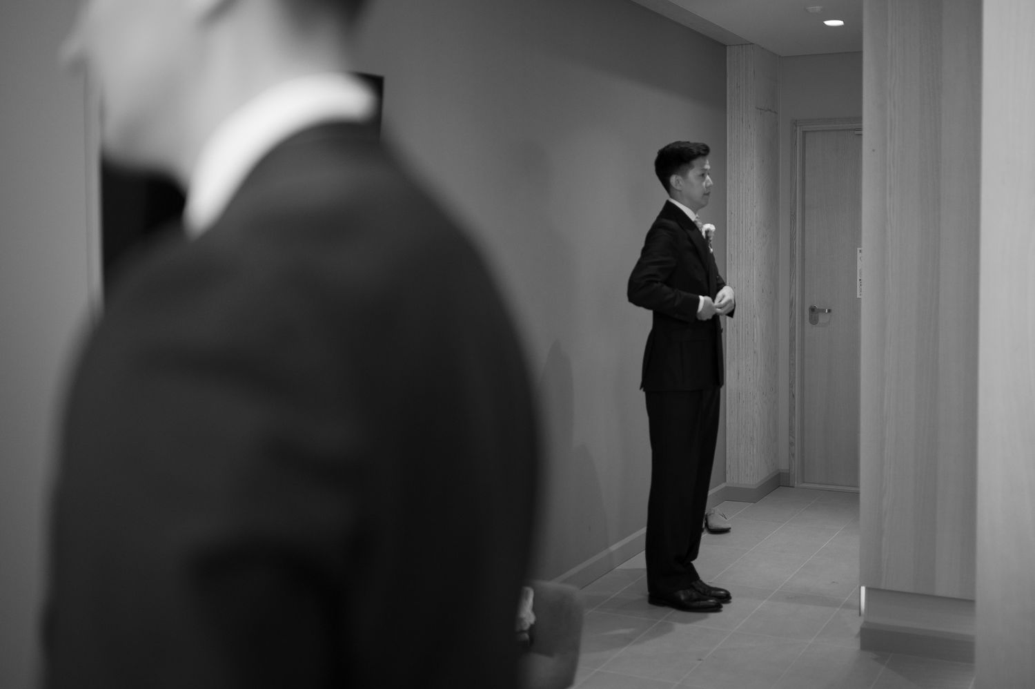 Black and white photo of a groom in a suit adjusting his jacket in a hallway before his wedding ceremony.