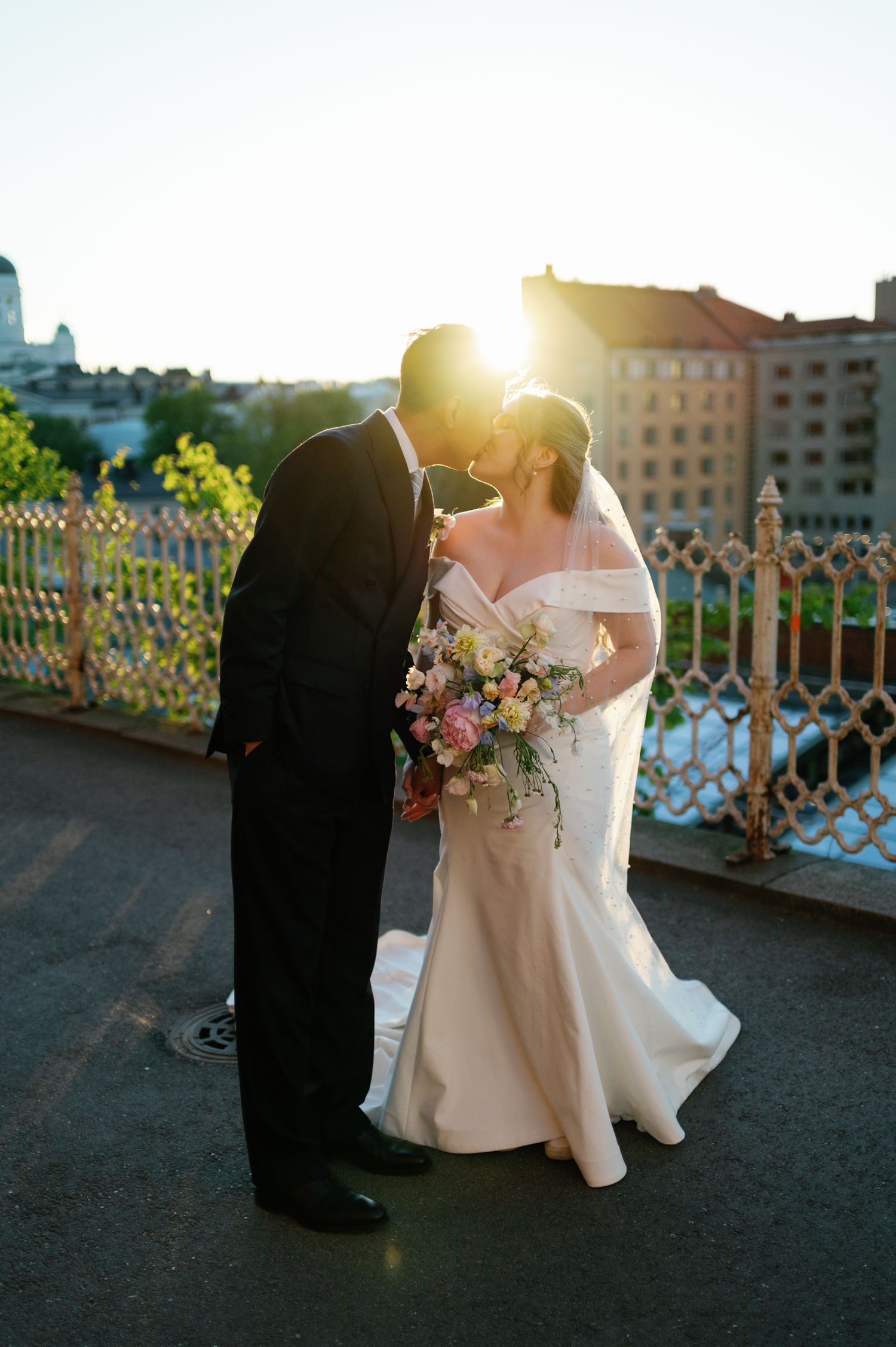 Bride and groom share a sunset kiss on a bridge, bride holding a colorful bouquet in a white off-shoulder gown.