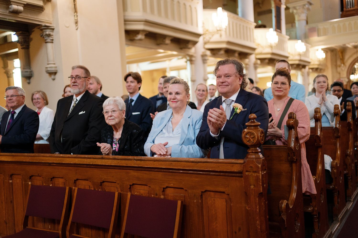 Wedding guests applauding in an ornate church, dressed in formal attire, seated in wooden pews during a ceremony.