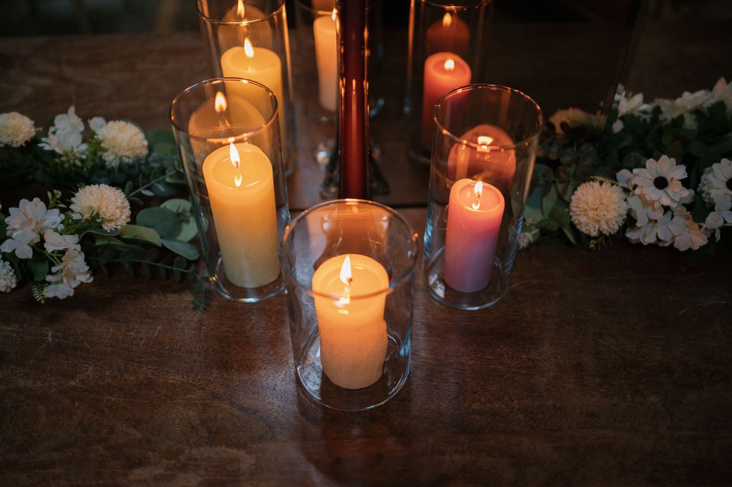 Lit white and pink candles in glass holders surrounded by white flowers and greenery on a wooden table.