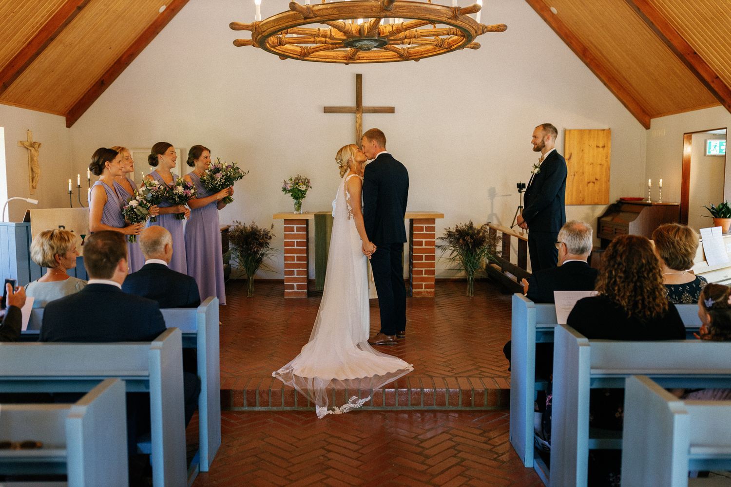 Interior of a chapel with exposed wooden beams, blue pews, and a cross mounted on the wall.