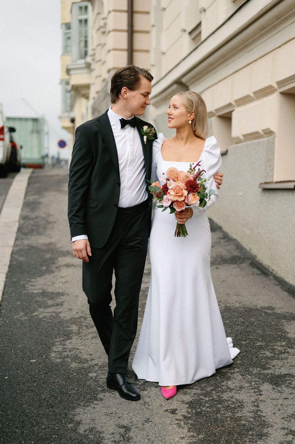 Wedding couple in formal attire posing on urban street with bride holding colorful bouquet.