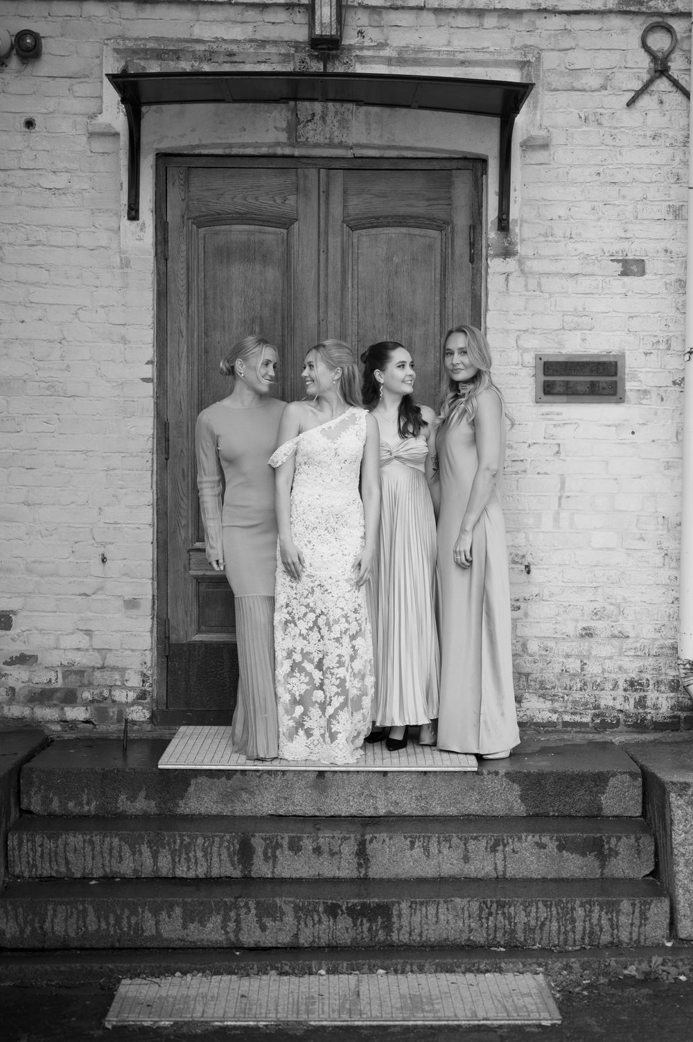 Black and white photo of wedding party members standing on a concrete platform against weathered brick wall.