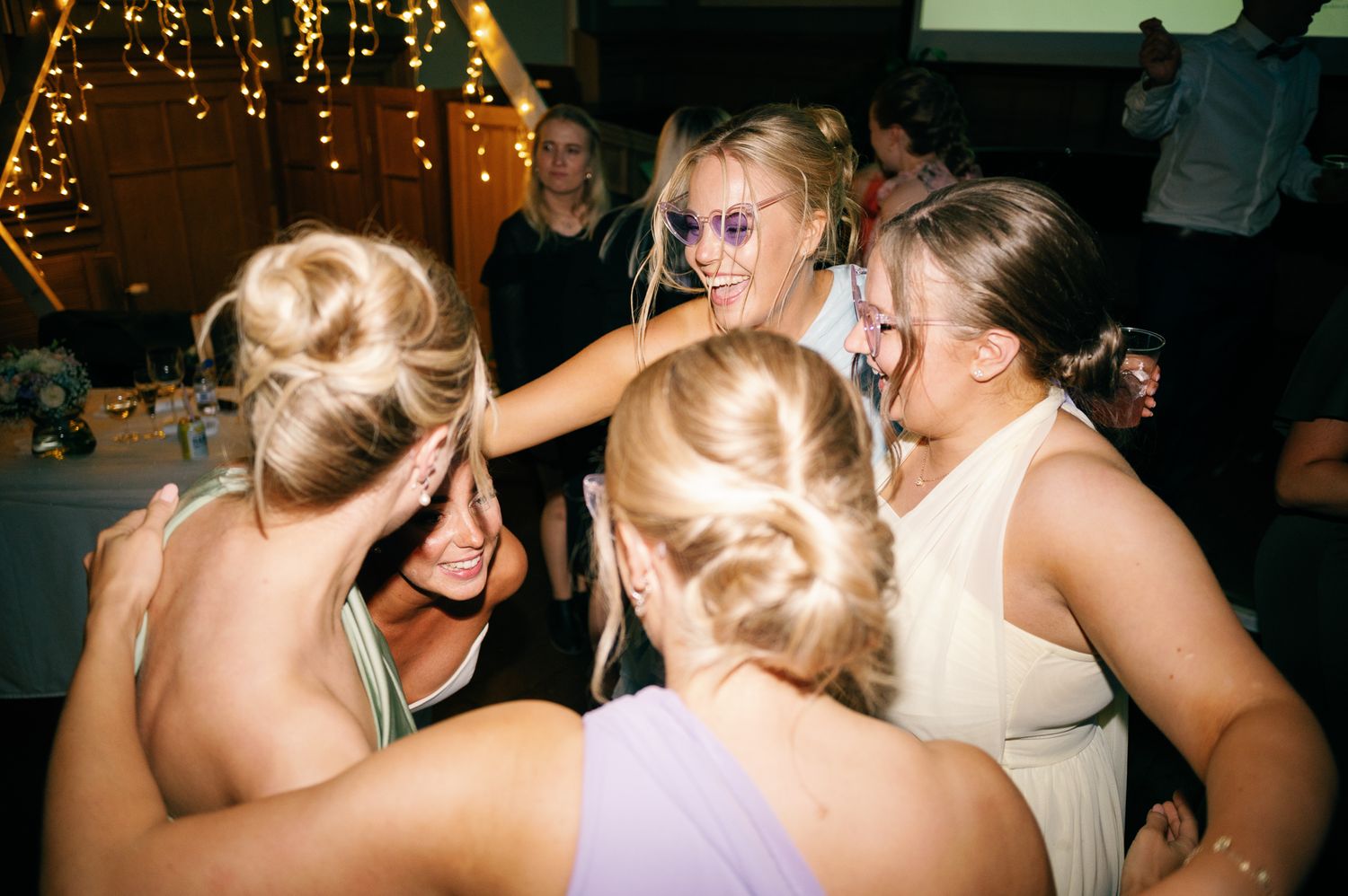 A group of wedding guests dancing and celebrating together on a dimly lit dance floor with festive lights in the background.