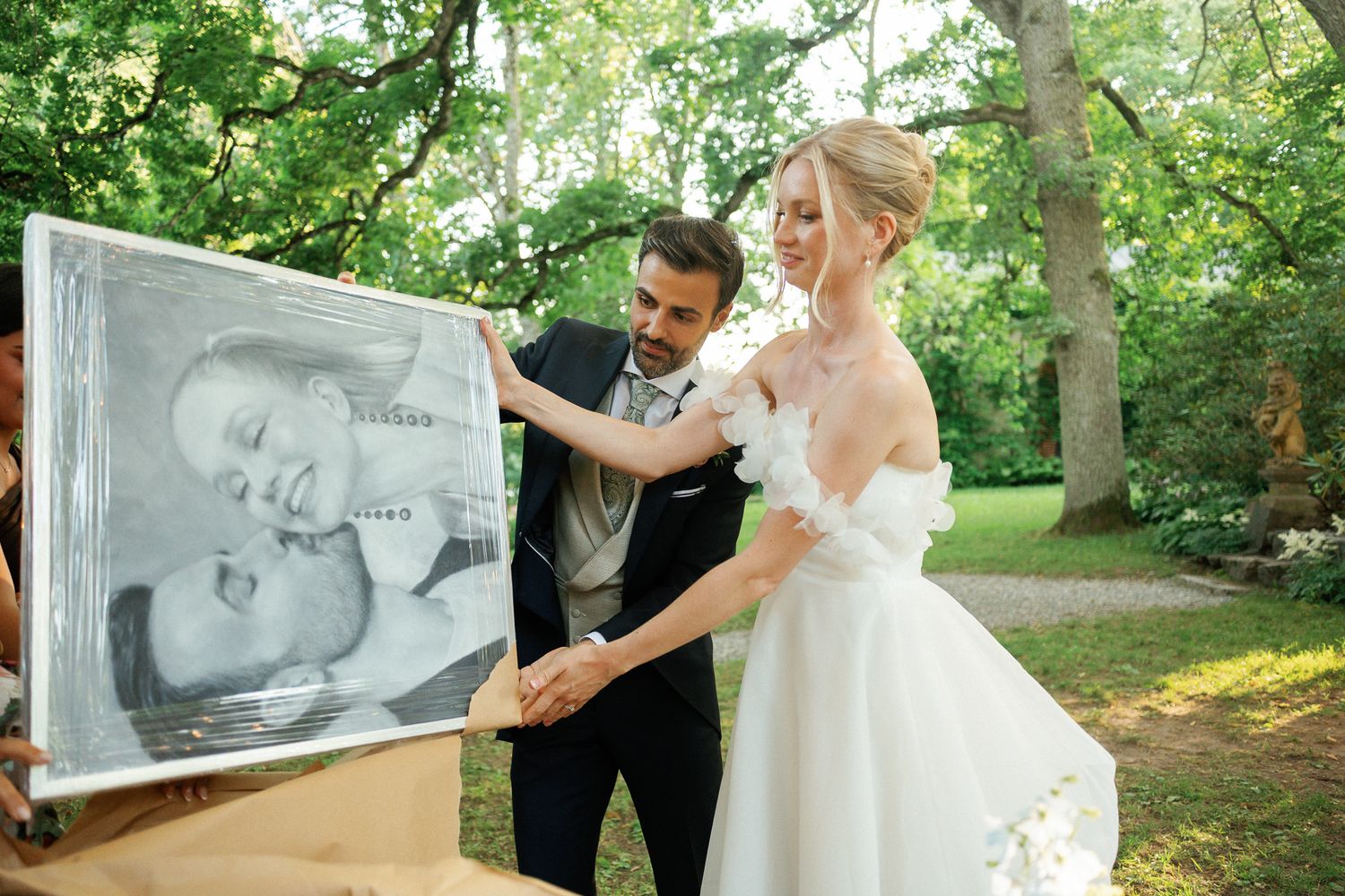 Bride and groom examining a charcoal portrait painting at their outdoor wedding reception surrounded by lush green trees.