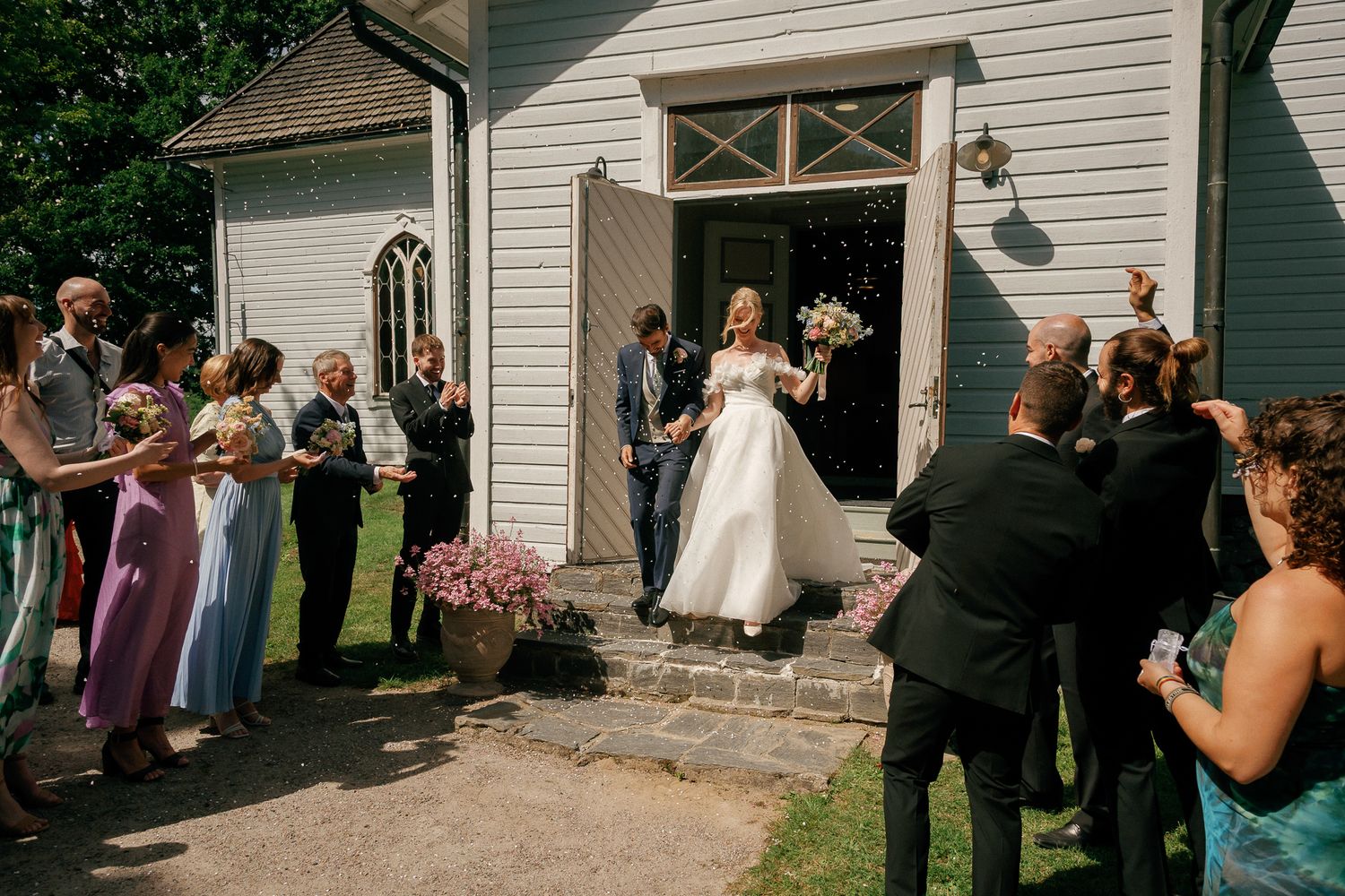 Bride exits a white chapel surrounded by smiling wedding guests outdoors on a sunny day.