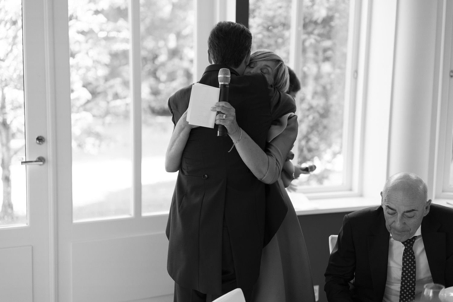 Two people share an emotional hug during a speech at a formal event, captured in black and white photography.