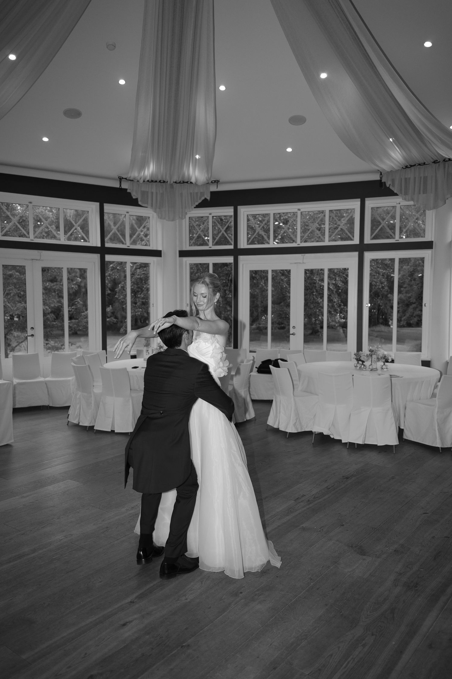 Bride and groom share their first dance in an elegant ballroom with draped ceilings and large windows.