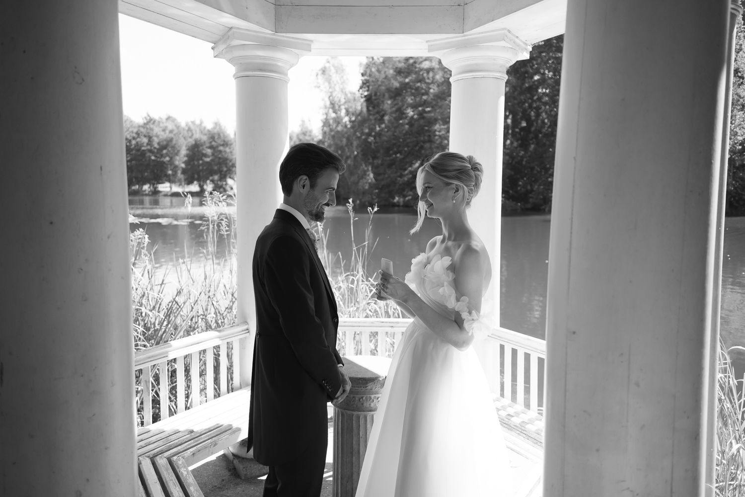 Black and white photo of a bride and groom exchanging vows in an elegant gazebo by a serene lake.