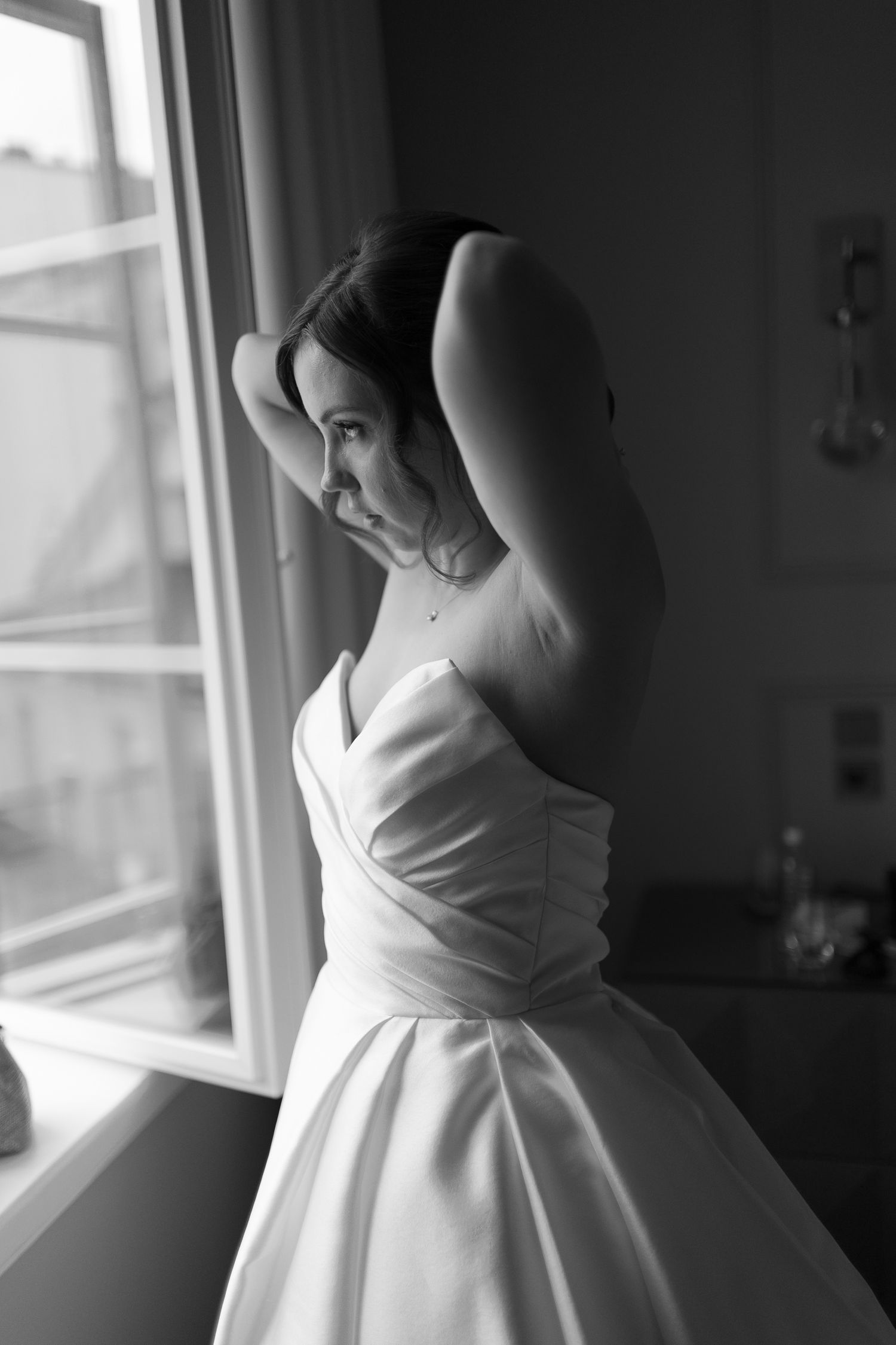 Elegant bride in strapless satin wedding dress posing by window in black and white portrait photo.