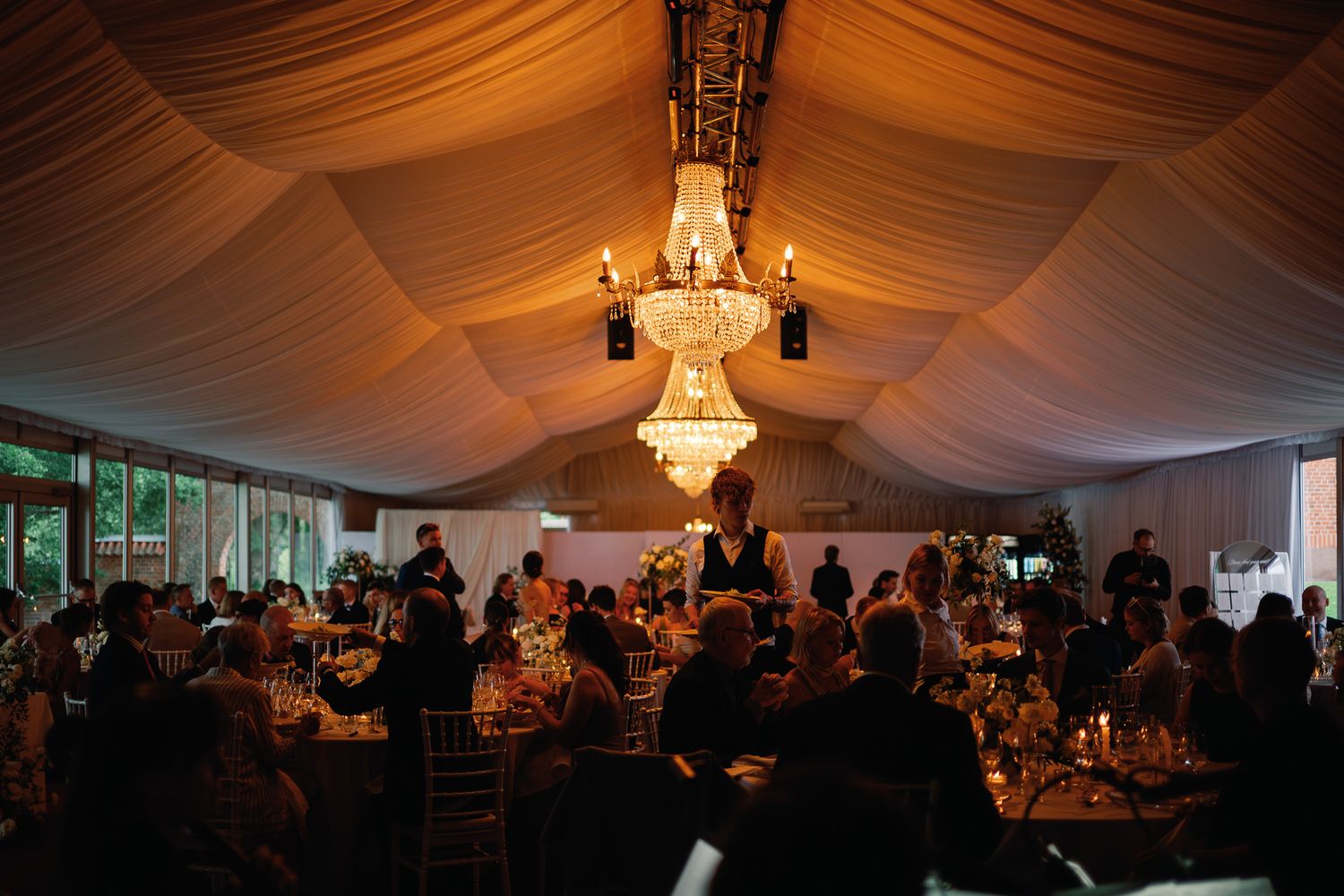 Elegant wedding reception under a draped tent with crystal chandelier, candlelit tables, and guests dining in warm golden light.