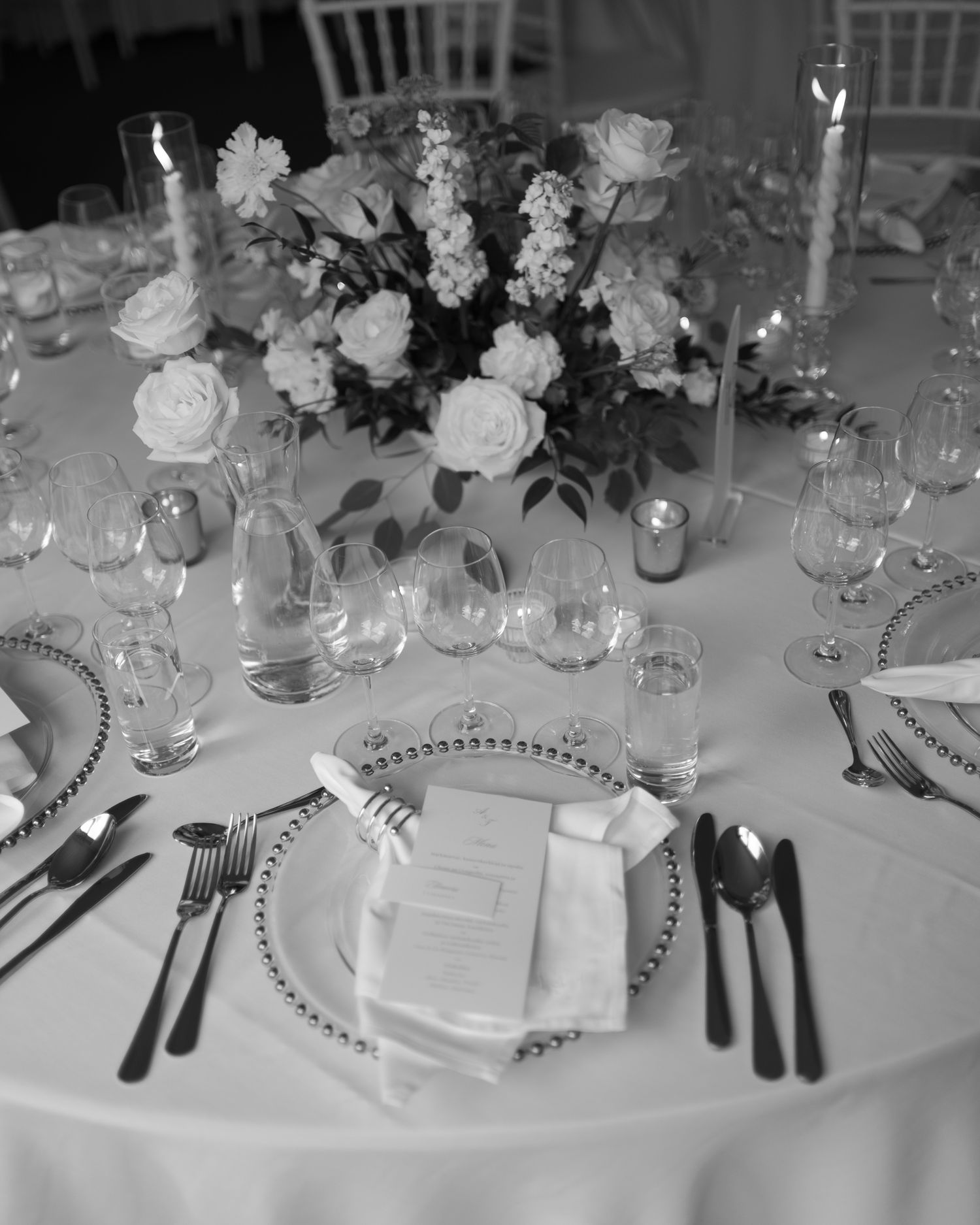 Black and white photo of an elegant wedding reception table with floral centerpiece, candles, crystal glasses, and formal place settings.