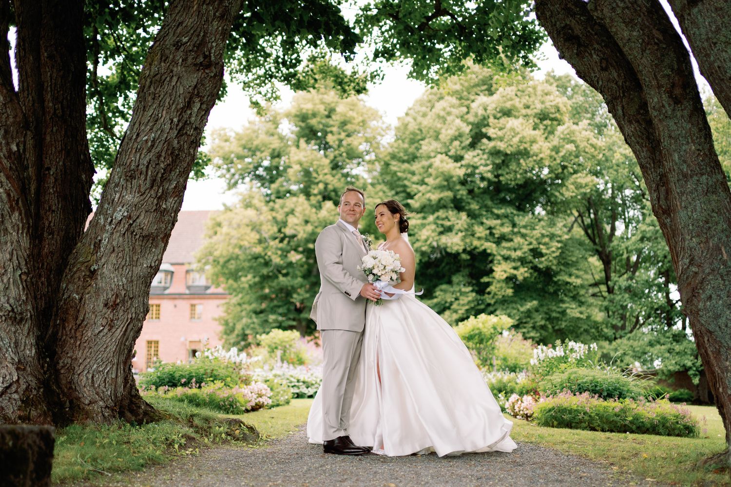 Bride and groom share a romantic moment under large trees in a lush garden setting on their wedding day.