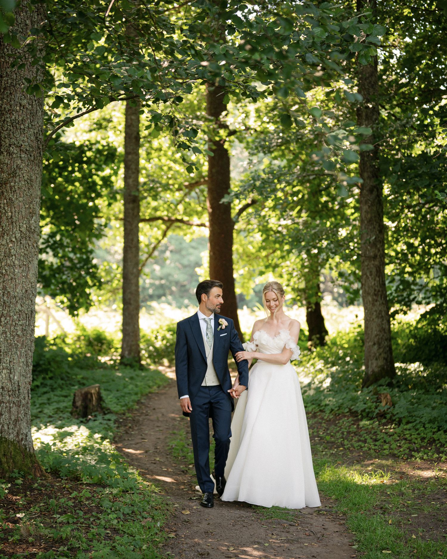 Bride and groom walk hand in hand along a sunlit woodland path, surrounded by tall green trees.