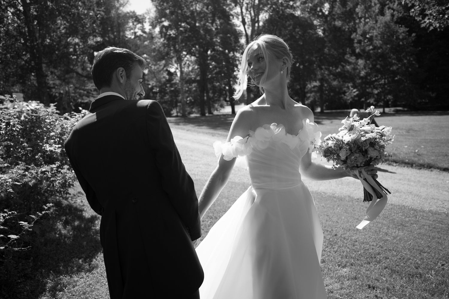 Black and white photo of a bride and groom holding hands walking through a lush park, bride holding a bouquet.
