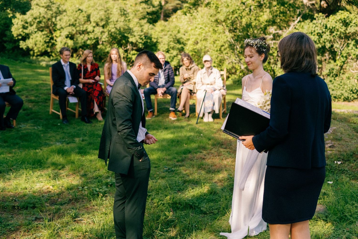 Wedding guests gather on a sunlit lawn for an outdoor ceremony near a grand yellow building.