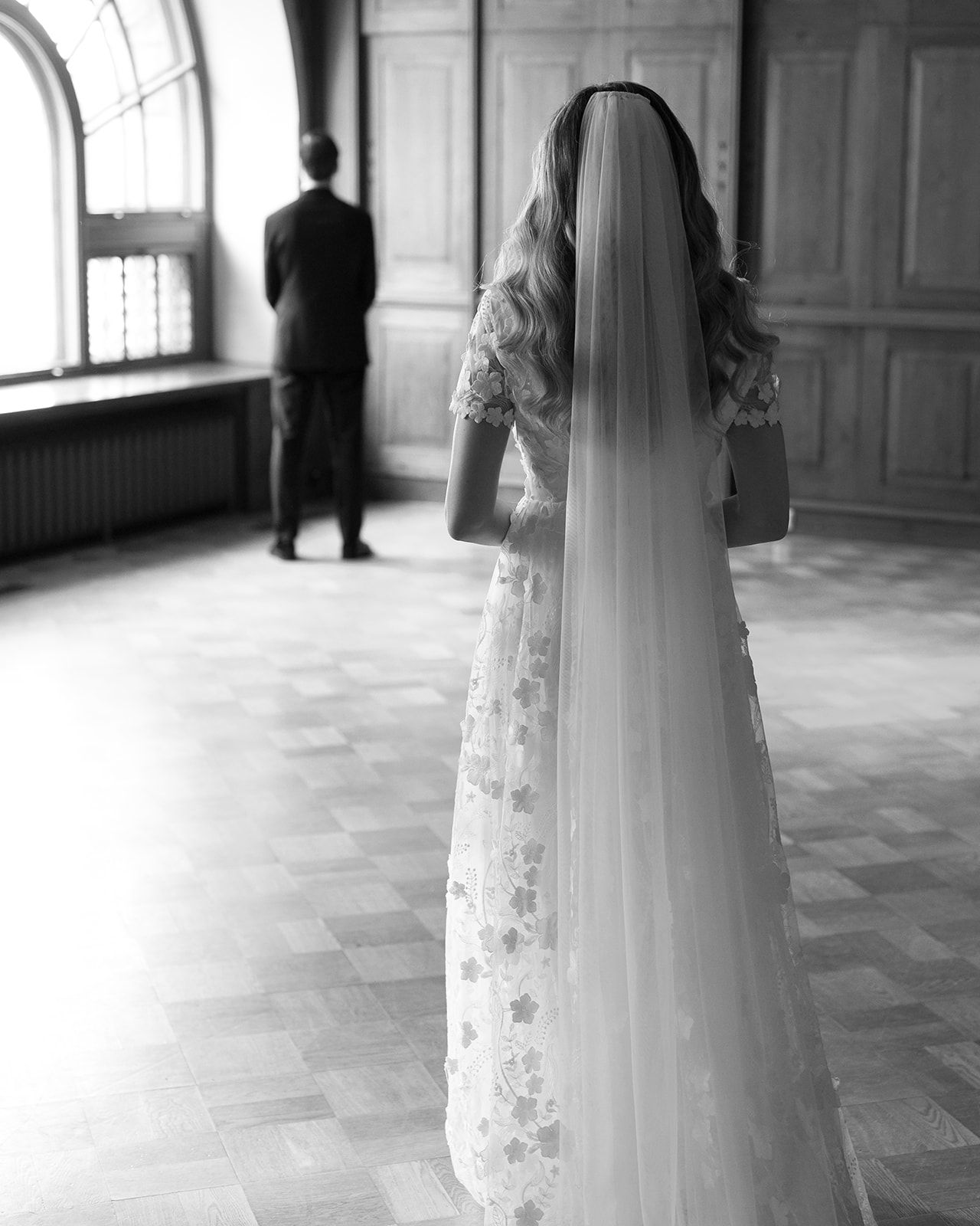 Black and white photo of a bride in a lace dress and long veil looking toward a silhouetted figure.