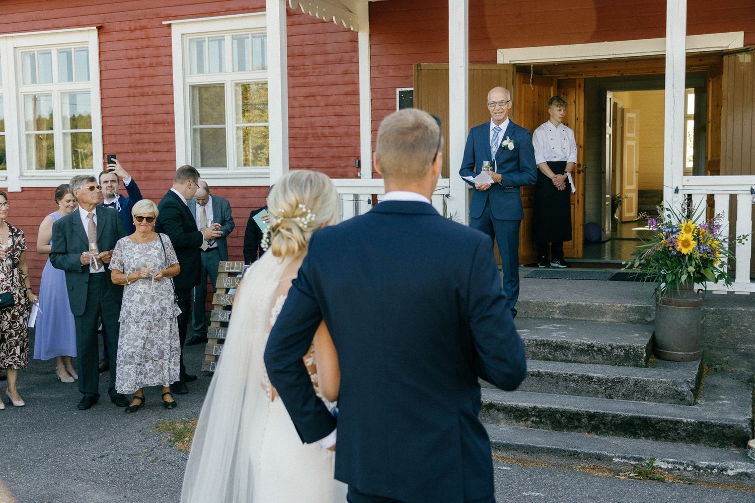 Guests mingle outside red wooden building during wedding celebration.