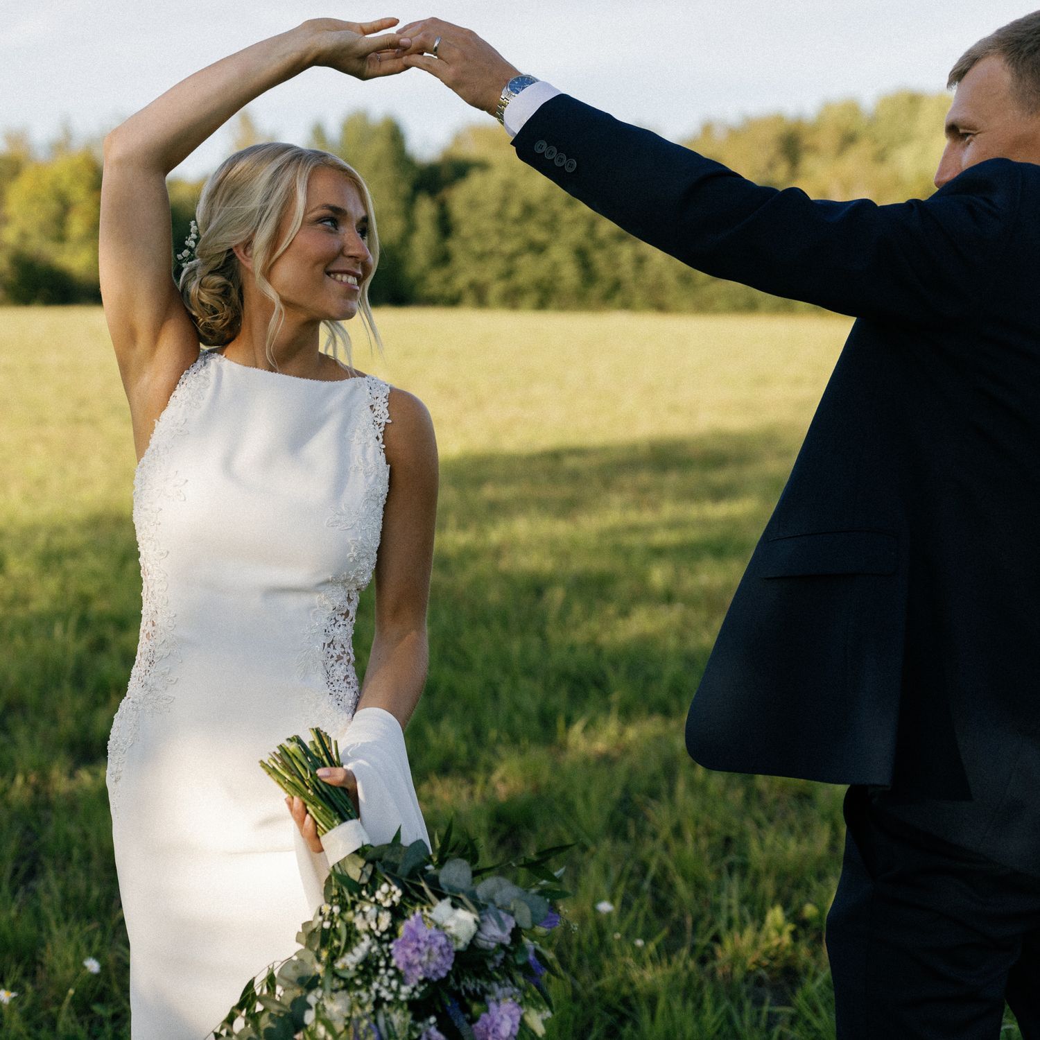 Joyful couple dancing in sunlit field with bride holding colorful bouquet.