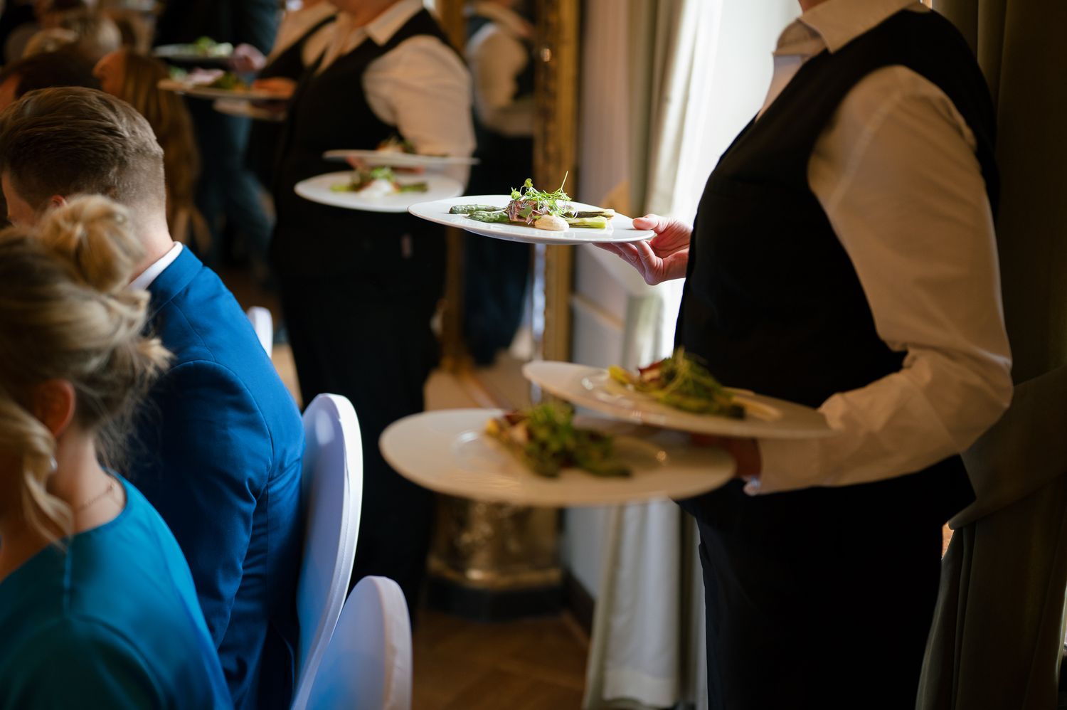 Restaurant server in black vest carries plates of food to diners seated at tables in an elegant dining room.