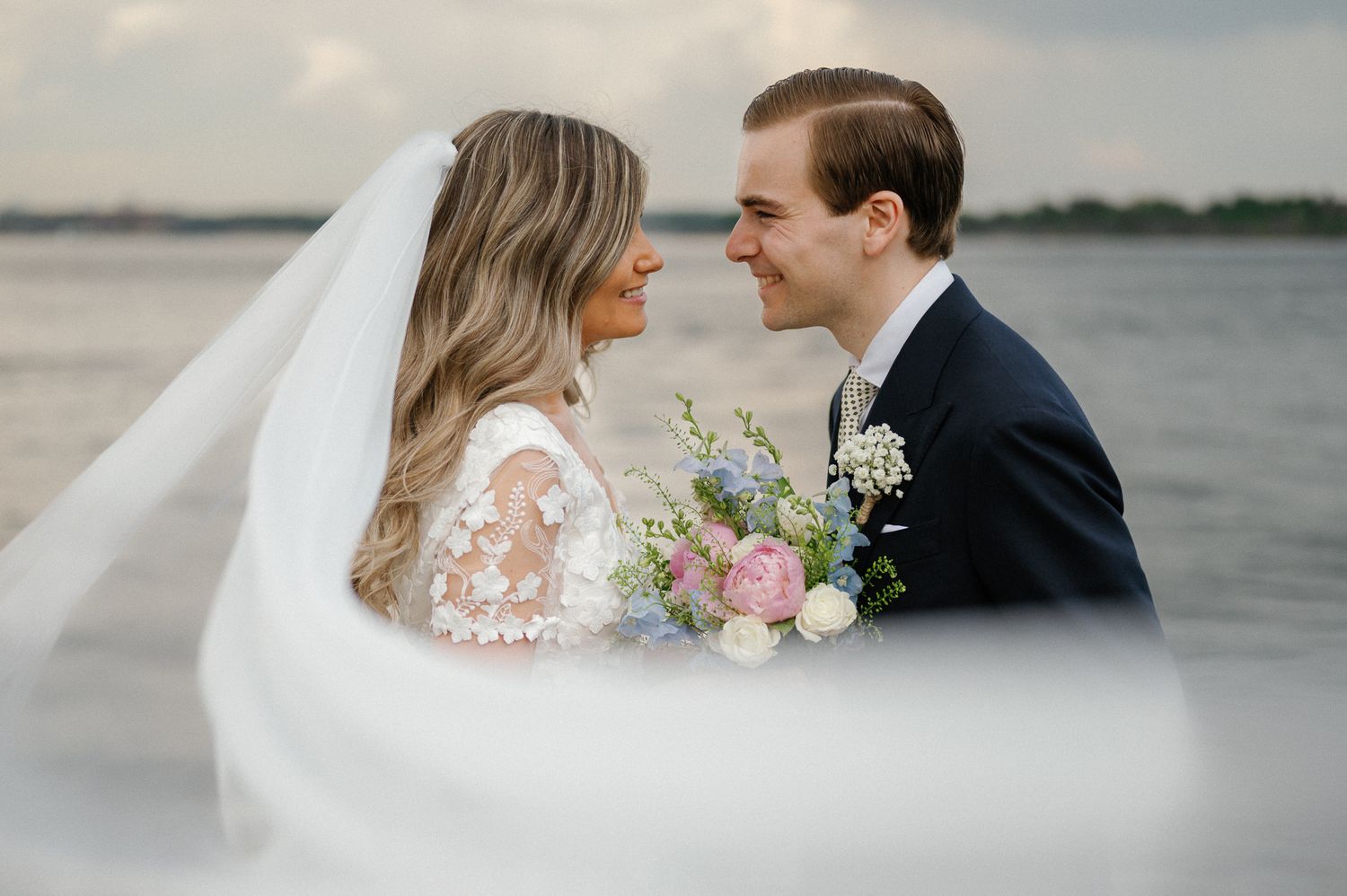 A romantic wedding couple shares tender moments by the water as a veil flows in the breeze.