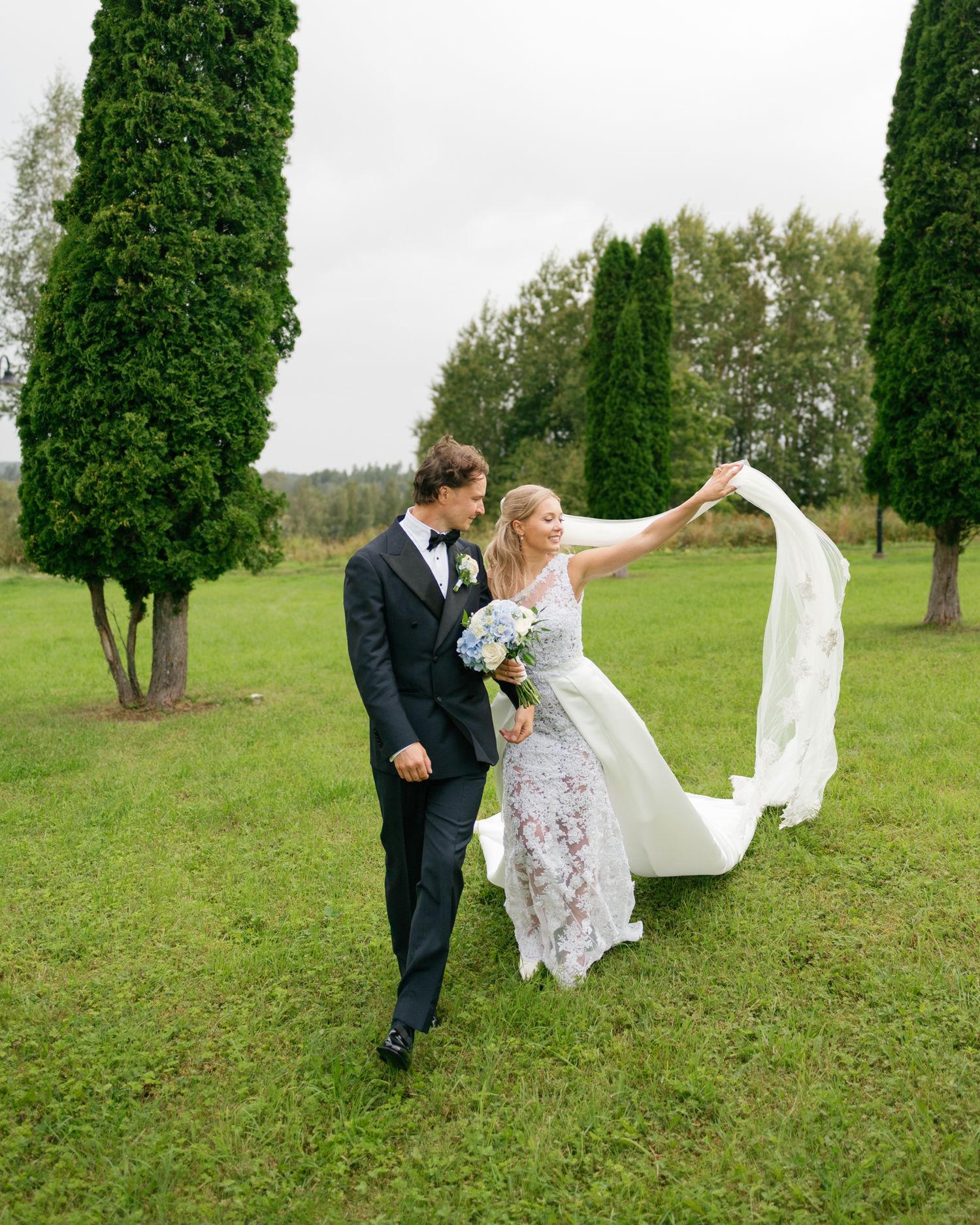 A couple in wedding attire playfully pose with a flowing veil in a green meadow with trees in the background.