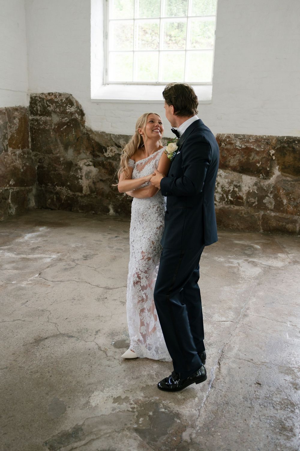A couple in formal attire dance together in an industrial space with white walls and stone features.