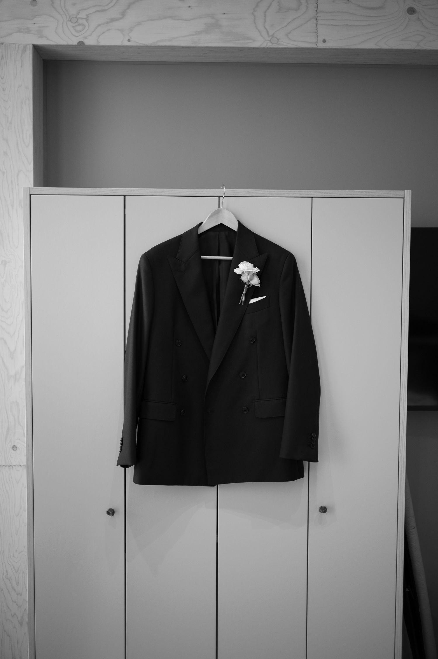 Black suit jacket with white boutonniere on hanger displayed against white cabinet doors in black and white photo.