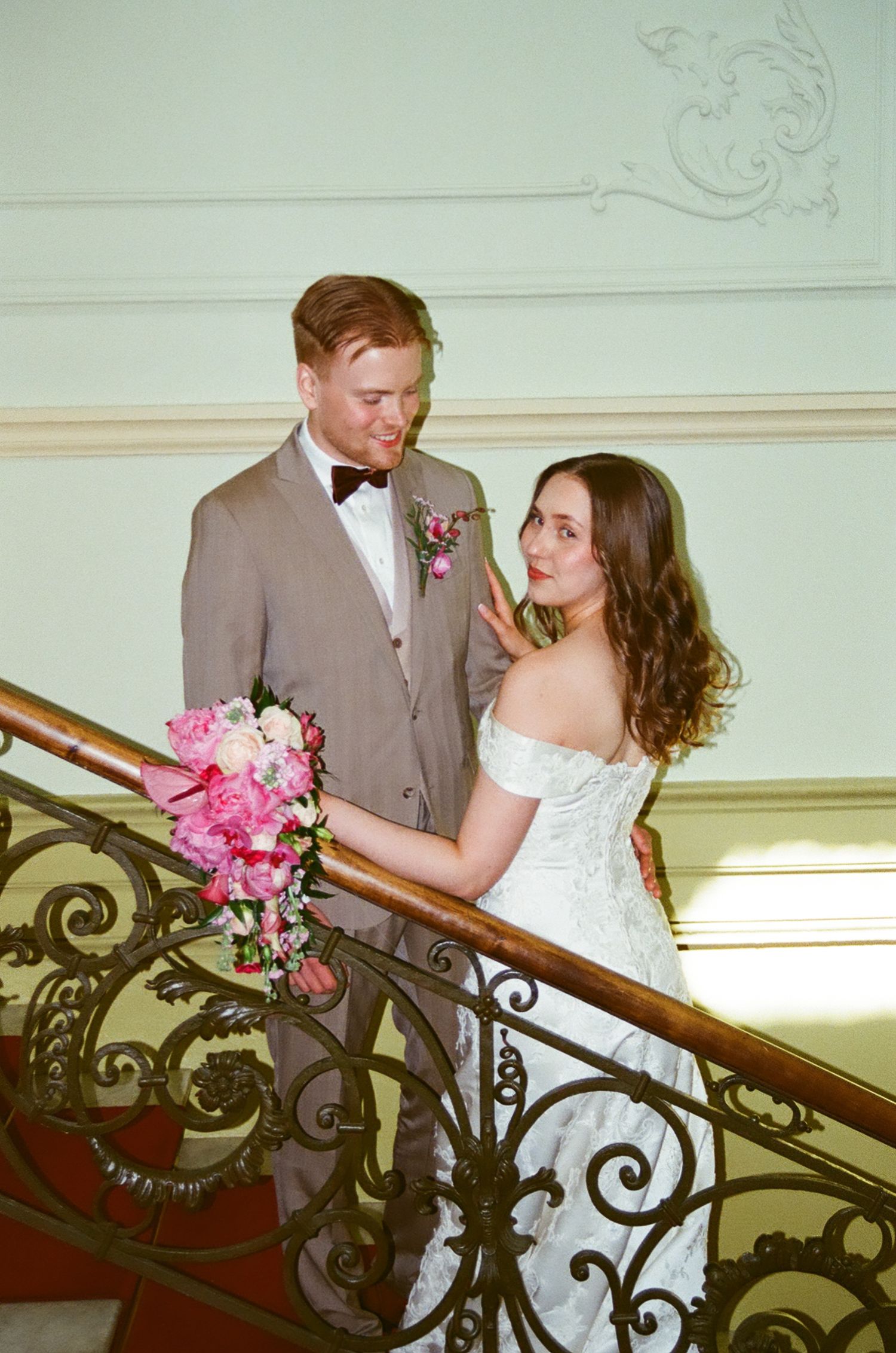 Bride and groom pose on ornate staircase, she holds pink peony bouquet, he wears gray suit with bow tie.