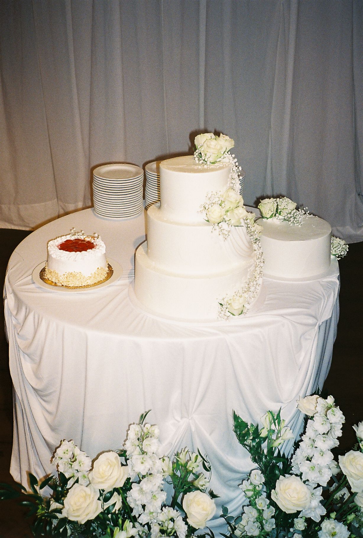 Elegant three-tier white wedding cake with floral cascades on draped table, surrounded by white roses and baby's breath.