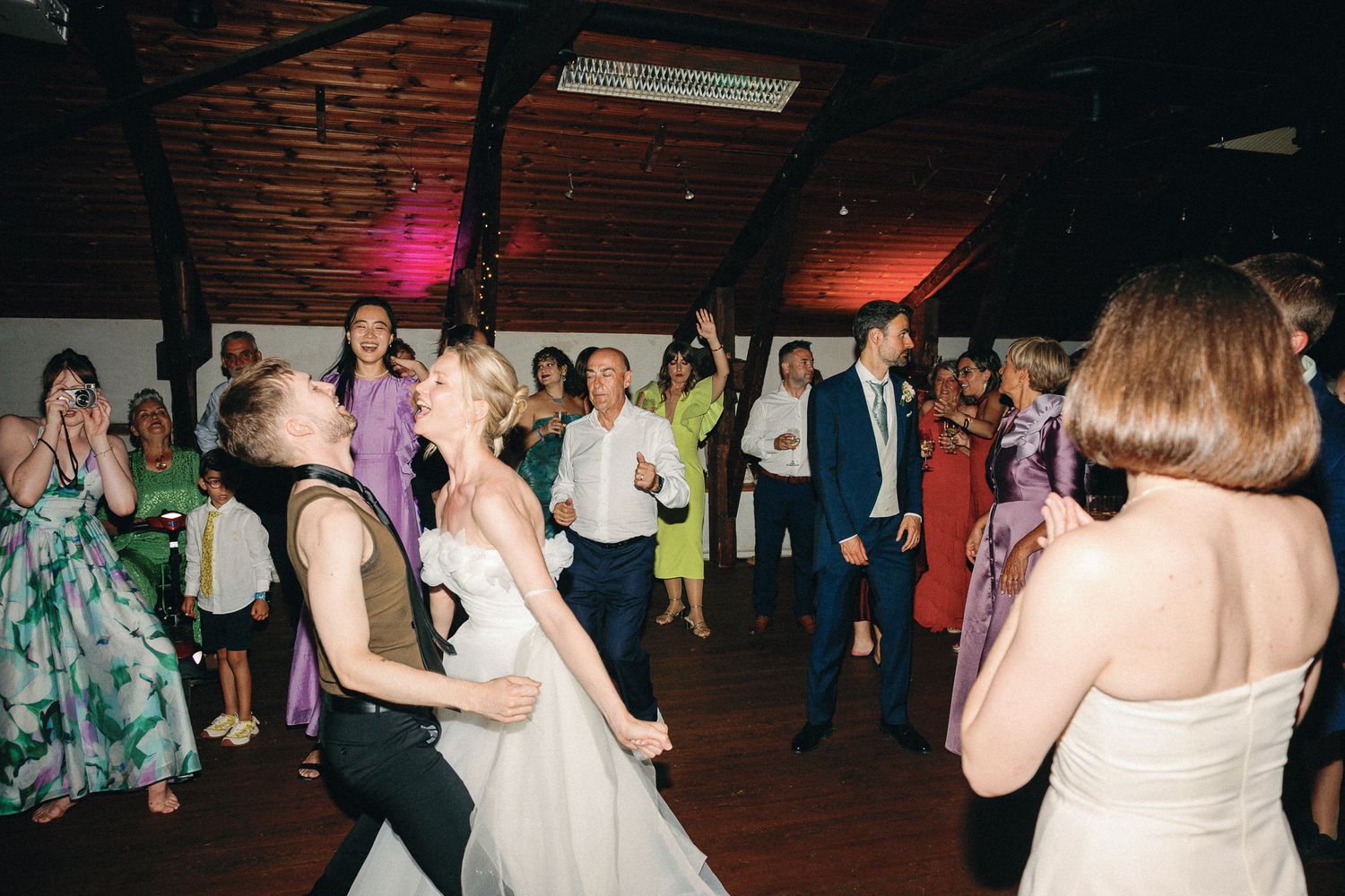 Bride and wedding guests laughing and dancing together at a lively indoor wedding reception.