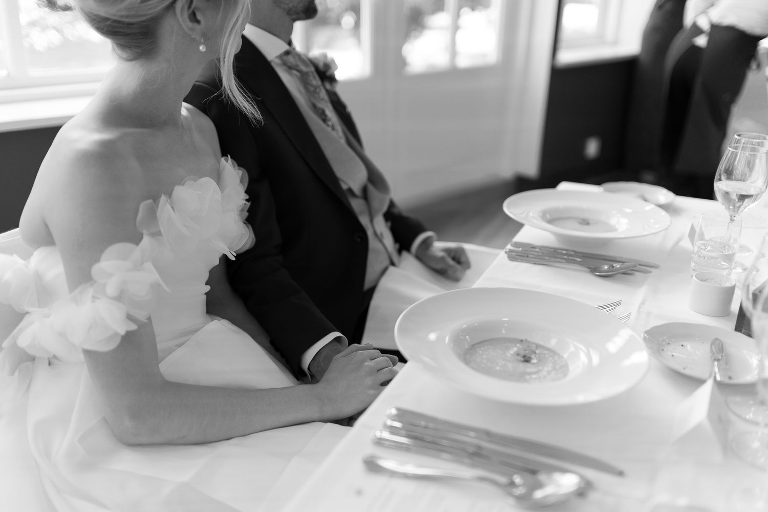 Black and white photo of bride and groom seated at elegant wedding reception table with fine china place settings.