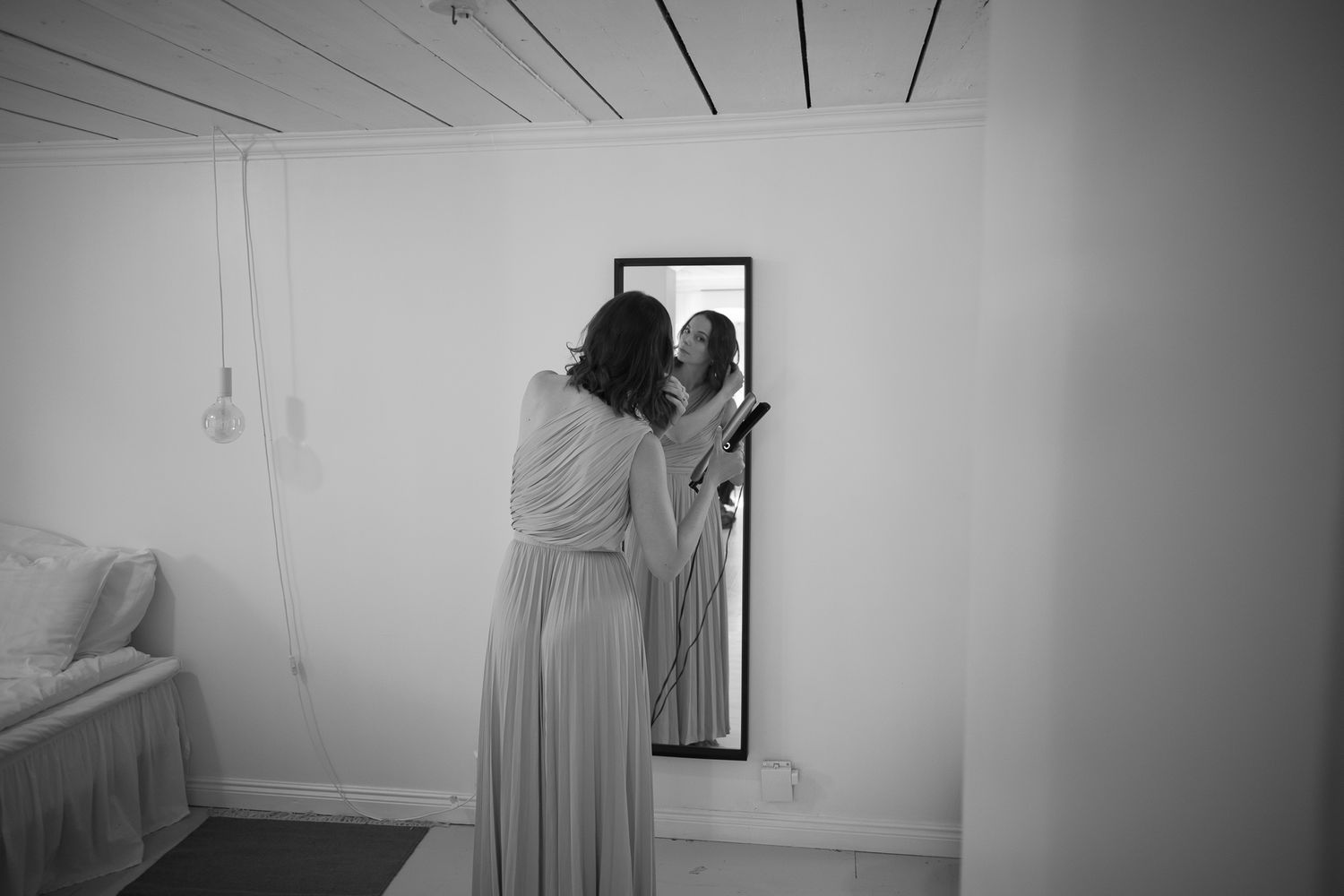 Black and white photo of a bride in a flowing gown examining her reflection in a tall mirror before her wedding.