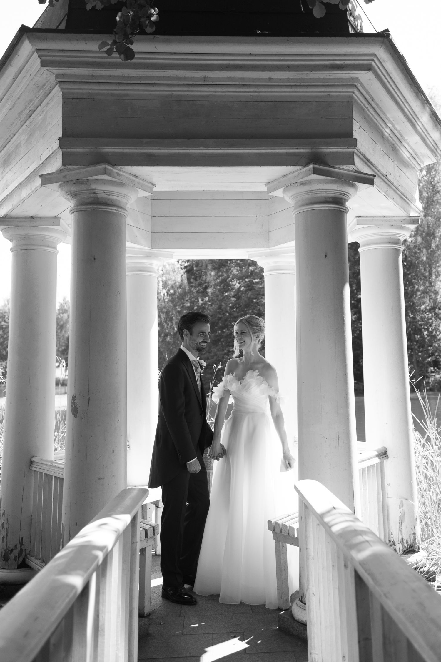 Black and white photo of a bride and groom holding hands in a classic white gazebo with columns on their wedding day.