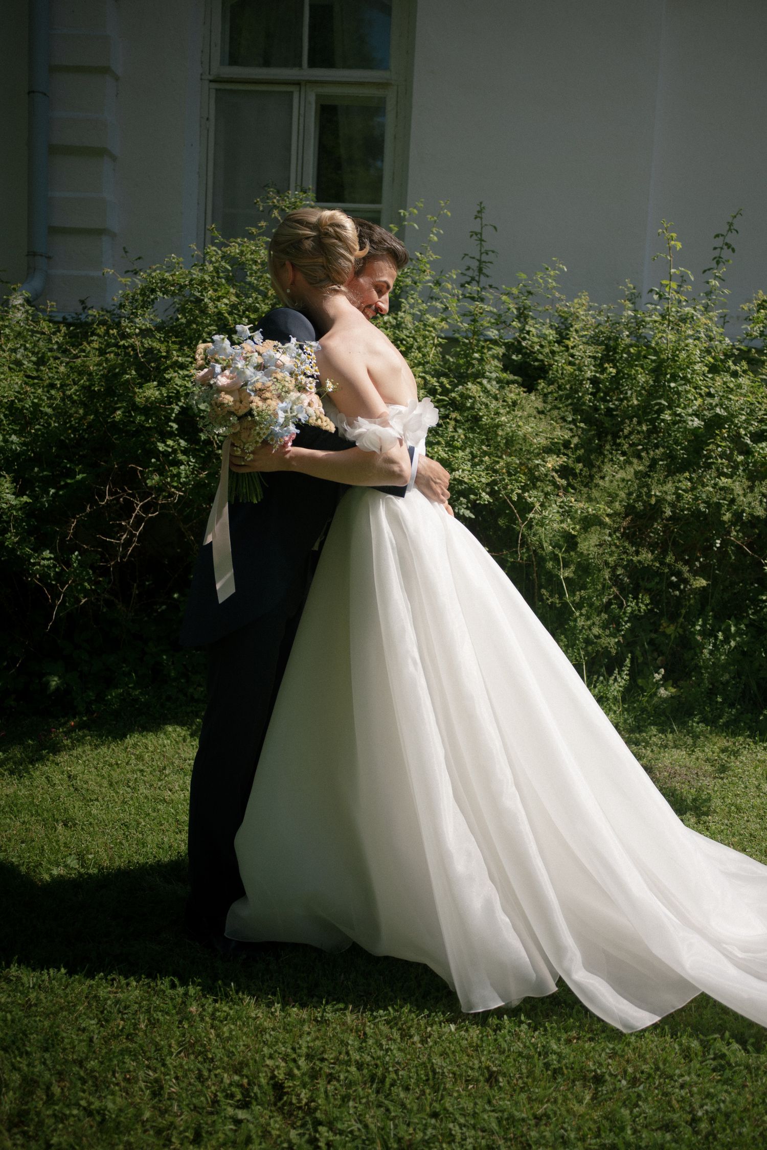 A bride in a white ball gown embraces her groom in a dark suit outdoors, holding a floral bouquet near green bushes.